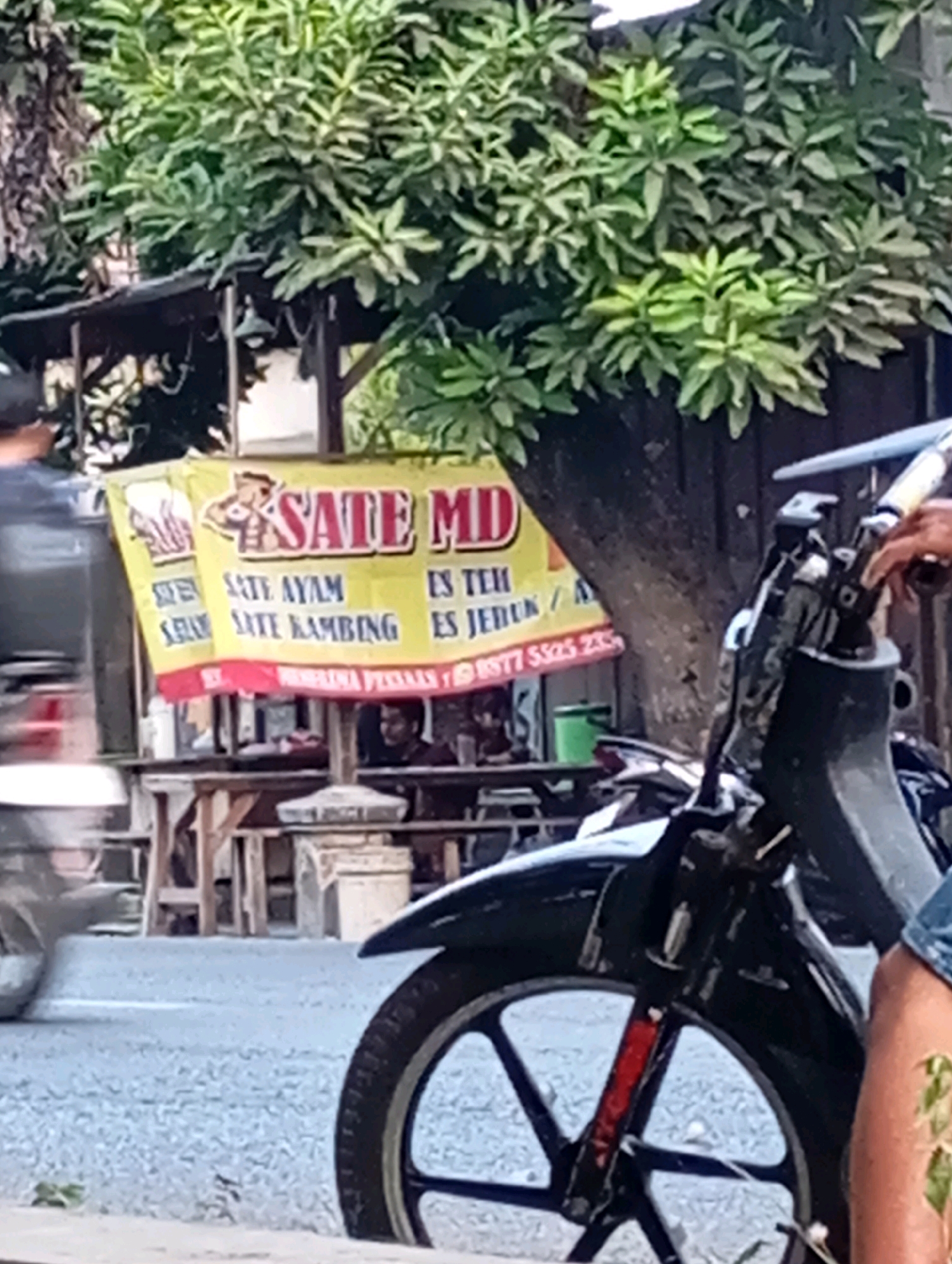 In Ketegan, Indonesia, a yellow banner advertises "SATE MD," featuring illustrations of muscular figures and listing menu items like "Sate Ayam" (chicken satay), "Sate Kambing" (goat satay), "Es Teh" (iced tea), and "Es Jeruk" (orange juice). The banner also displays a phone number, "0877 5525 2350." The banner is displayed in front of a food stall with a wooden structure. A person on a black motorcycle is partially visible in the foreground, with its front wheel and leg of the rider in view. The scene is set outdoors with a tree providing shade.