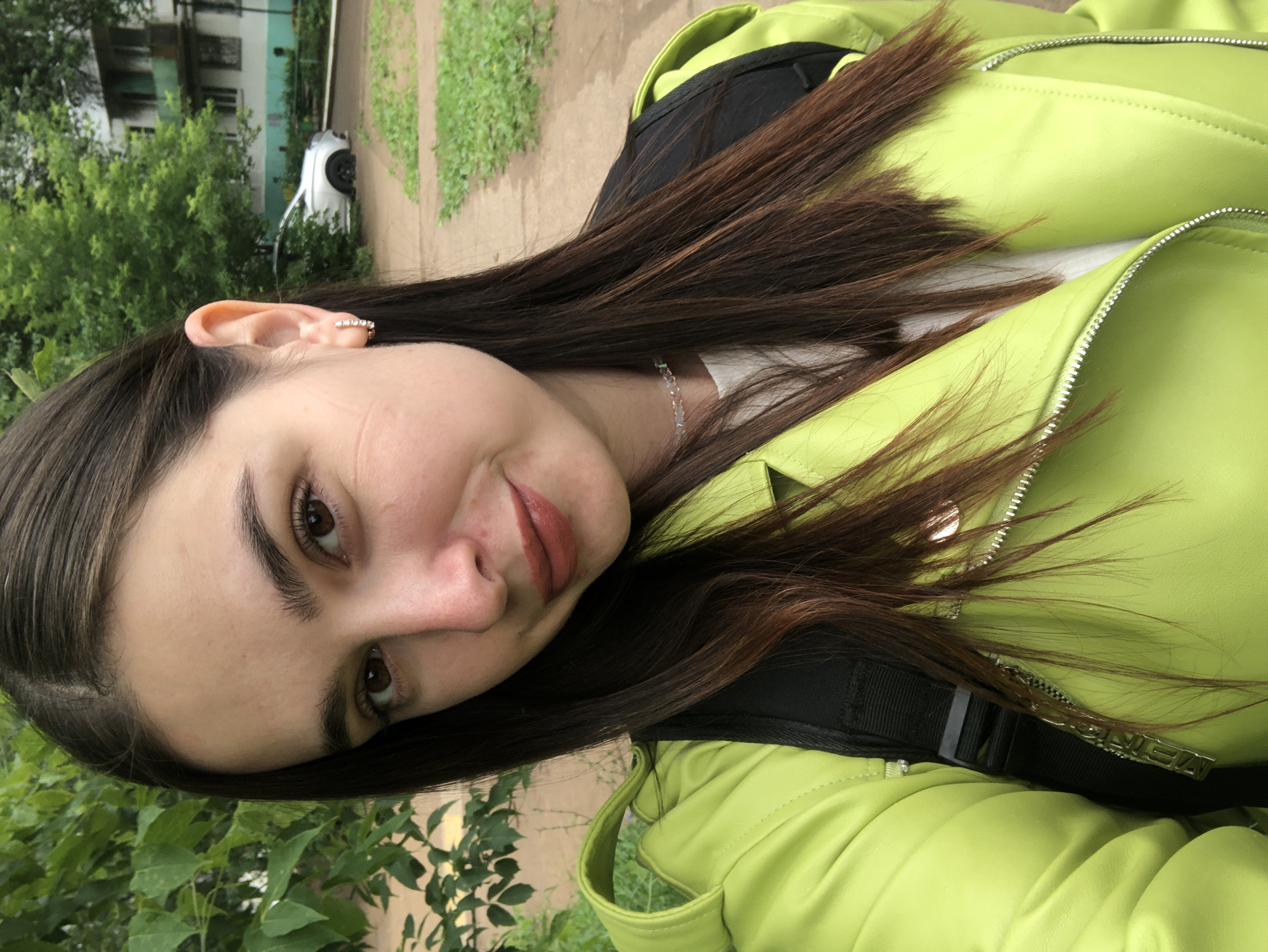 A young woman with long, dark brown hair takes a selfie outdoors. She is wearing a bright lime green jacket with a black backpack. A silver necklace with small charms is visible around her neck. She has a light dusting of freckles across her nose and cheeks and is wearing a natural-looking lip color. Her right ear is adorned with a small, sparkly earring. The background shows a pathway, lush green foliage, and a building in the distance. A white car is parked on the side of the road. The lighting suggests it is daytime, and the greenery indicates a mild climate. The overall impression is casual and relaxed, with the woman smiling slightly at the camera.