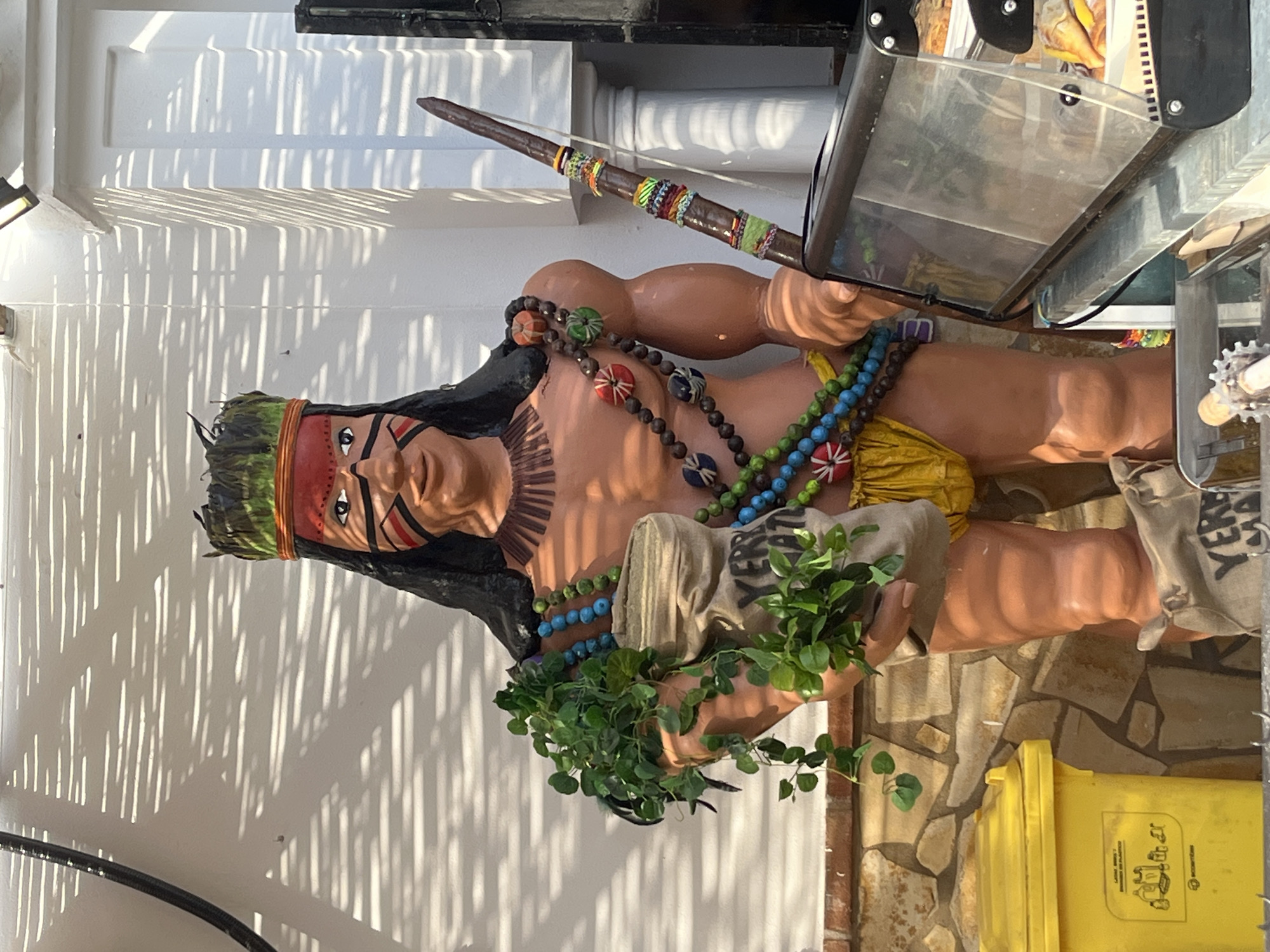 The image shows an upright, life-sized statue of a male figure, resembling an indigenous person, positioned outdoors under bright sunlight in what appears to be Fuengirola, Spain. The statue has dark hair, a red and green headband, and distinctive red, black, and white face paint. It is dressed in a yellow loincloth and adorned with multiple colorful beaded necklaces in shades of green, blue, brown, red, and white.
In its right hand, the figure holds a decorated spear or staff wrapped with colorful bands. Its left hand cradles a beige, sack-like object from which vibrant green artificial foliage emerges. The statue also appears to be holding a display case or sign holder, partially visible on the right.
The background features a bright white wall with prominent, elongated parallel shadows cast by strong sunlight, suggesting either morning or late afternoon. The ground is paved with irregular stone tiles. A yellow recycling bin is visible in the lower right, bearing the text "LARGE RECYCLED PACKAGING" and "ENVASES GRANDES RECICLABLES," along with recycling symbols. The statue serves as a decorative element, likely for a commercial establishment in a sunny, public setting.