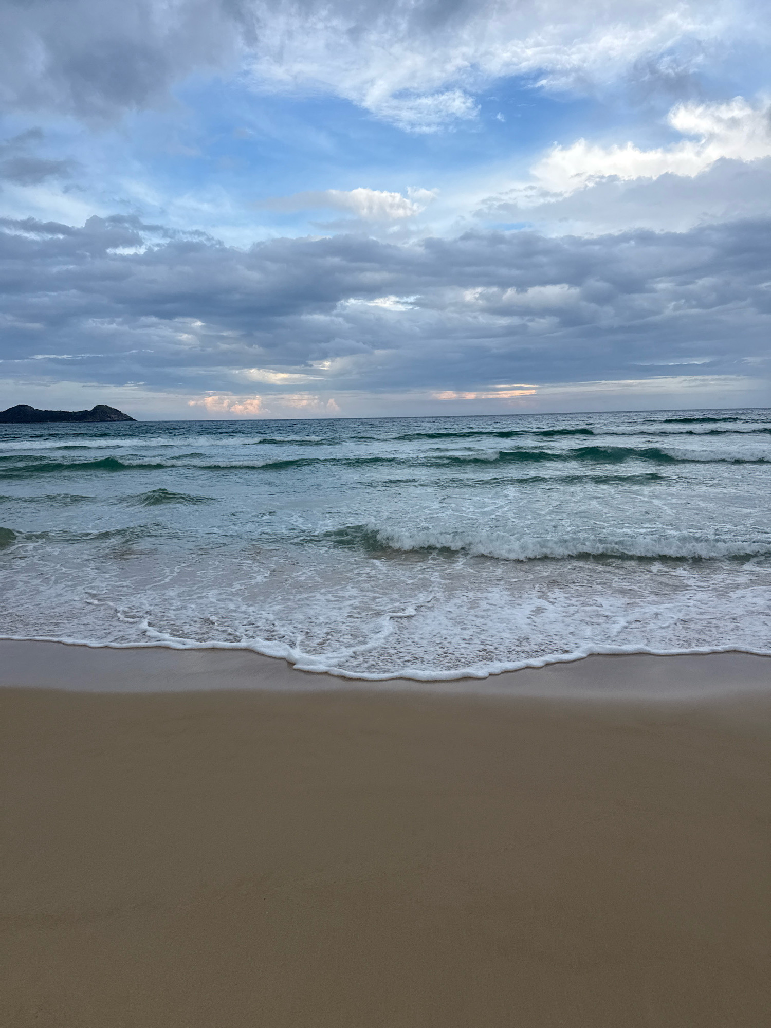 The image shows a beach scene with a sandy shore in the foreground and the ocean extending to the horizon. Waves are breaking on the shore, creating white foam. In the distance, a dark, hilly landmass is visible against a cloudy sky. The sky is filled with various shades of gray and blue clouds, with some hints of light breaking through. The water appears to be a greenish-blue color. The image is a still shot, with no people or specific structures visible, suggesting a natural coastal environment.