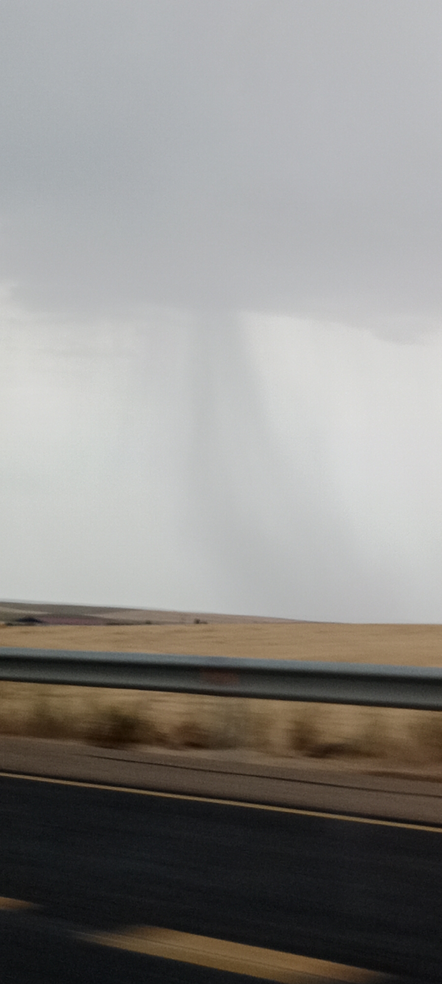 The image captures a dynamic weather phenomenon over a rural landscape, likely in the vicinity of Cerecinos de Campos, Spain. A distinct rain shaft, appearing as a grey, vertical streak, descends from a heavy, overcast sky. The sky itself is a uniform, pale grey, suggesting a cloudy or stormy day. Below the sky, a hazy band of lighter grey extends horizontally, potentially indicating a weather front or reduced visibility.
The middle ground features a vast expanse of dry, golden-brown terrain, likely agricultural fields. A horizontal, metallic guardrail runs across the lower portion of this area, suggesting the scene is viewed from a road or highway. The guardrail is dark grey with some lighter metallic reflections.
The foreground is dominated by a dark asphalt road, with two distinct yellow lines running parallel to each other. The road surface appears to be in motion blur, indicating the photograph was taken from a moving vehicle. The overall impression is one of a journey through a landscape under a dramatic sky, with the rain shaft being the focal point of the atmospheric conditions.