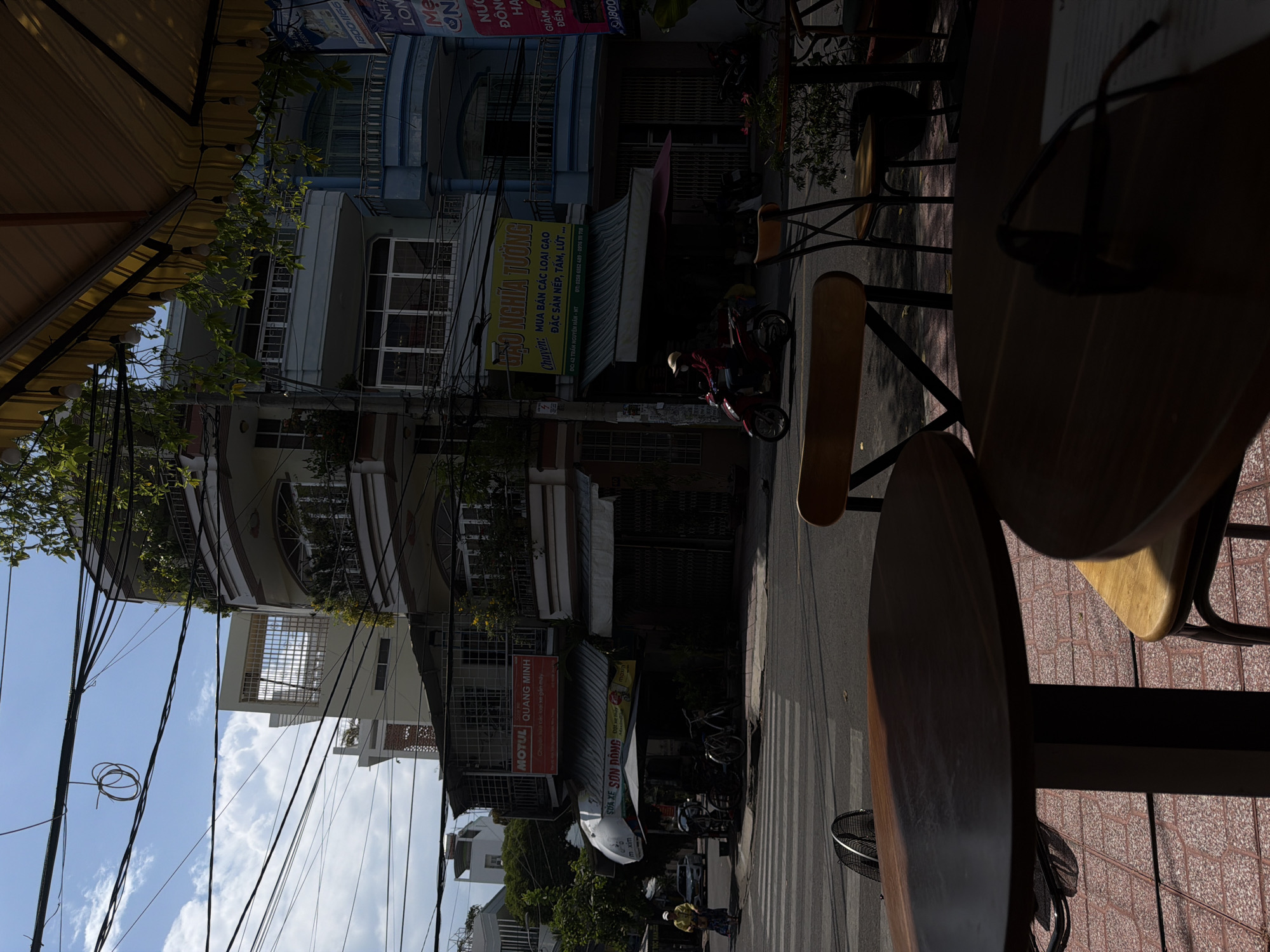 The scene is set on a street in Nha Trang Ward, Vietnam, with the perspective originating from beneath the awning of a storefront cafe. In the foreground are tables and chairs, one adorned with a pair of dark glasses. The street is lined with buildings, including one blue building with vertically oriented windows. Several signs and advertising are visible on the building. A motorcycle carrying two people travels down the street. A plethora of electrical wires cross the sky. The sky is blue and partly covered with clouds.