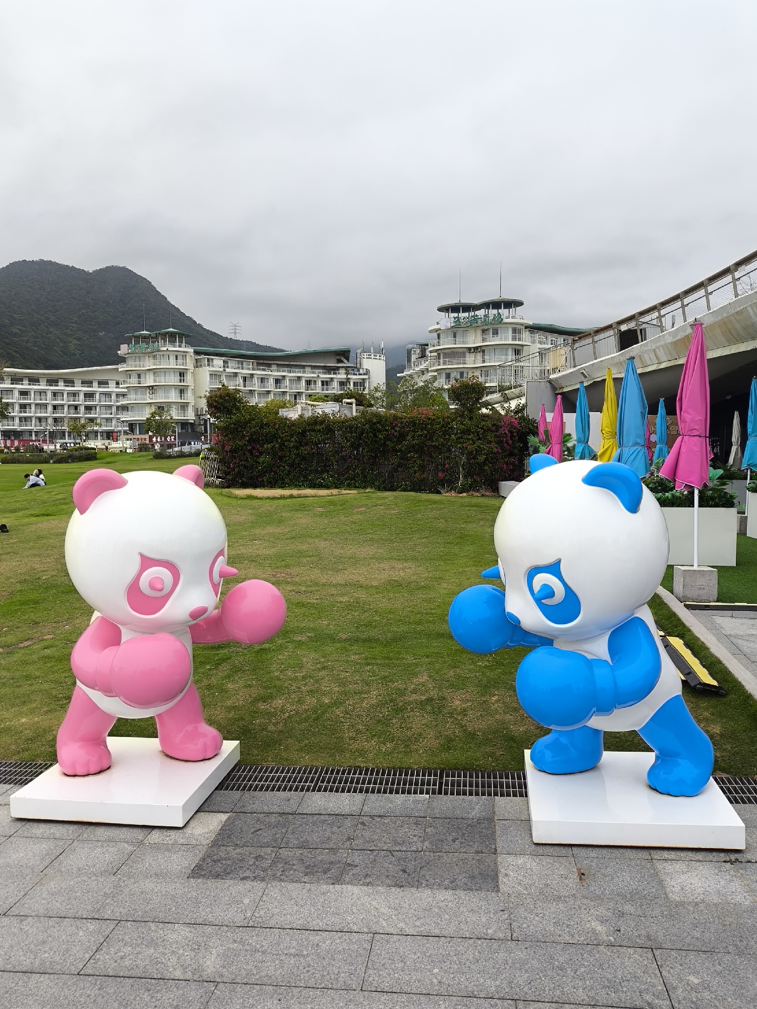 Two panda statues stand on a paved area with a green lawn behind them. One panda is pink, and the other is blue. Both are posed as if ready to box. Behind the pandas is a building with multiple stories and balconies, partially obscured by trees and bushes. To the right of the building, colorful umbrellas are folded and stored. In the background, a mountain is visible under a cloudy sky. This scene is located in the Yantian District, China.