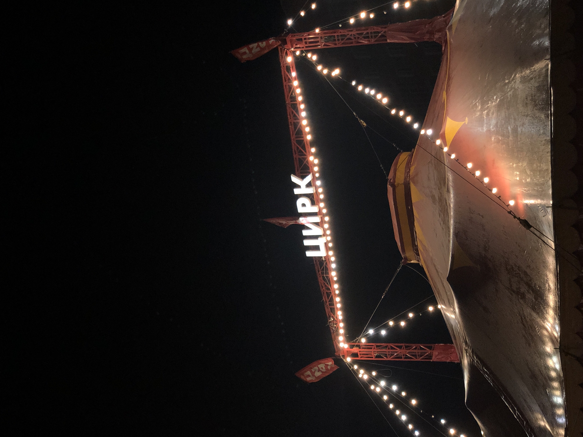 The image captures an evening scene at a circus tent in Petropavlovsk-Kamchatsky, Russia. The primary subject is the circus tent itself, a large, imposing structure with a metallic-looking surface reflecting the surrounding lights. A red, metallic support structure, adorned with numerous incandescent bulbs, extends from the left side of the frame towards the tent. These lights, strung along cables, create a festive and somewhat magical atmosphere, illuminating the dark night sky.
Prominently displayed on the support structure is the word "ЦИРК" (Tsirk), meaning "circus" in Russian, rendered in large, white, illuminated letters. The letters are placed vertically, running upwards along the structure. Below the main sign, a small red banner with some indistinct markings is also visible.
The time of day is clearly night, with the blackness of the sky providing a stark contrast to the illuminated elements of the circus. There are no visible people, suggesting the image focuses on the architectural and atmospheric aspects of the circus setup. The lighting from the bulbs and the tent's own illumination create a warm, inviting glow, hinting at excitement and entertainment. The weather appears clear, allowing the lights to shine brightly without obstruction. The overall impression is one of a classic circus setting, ready for a performance under the night sky.