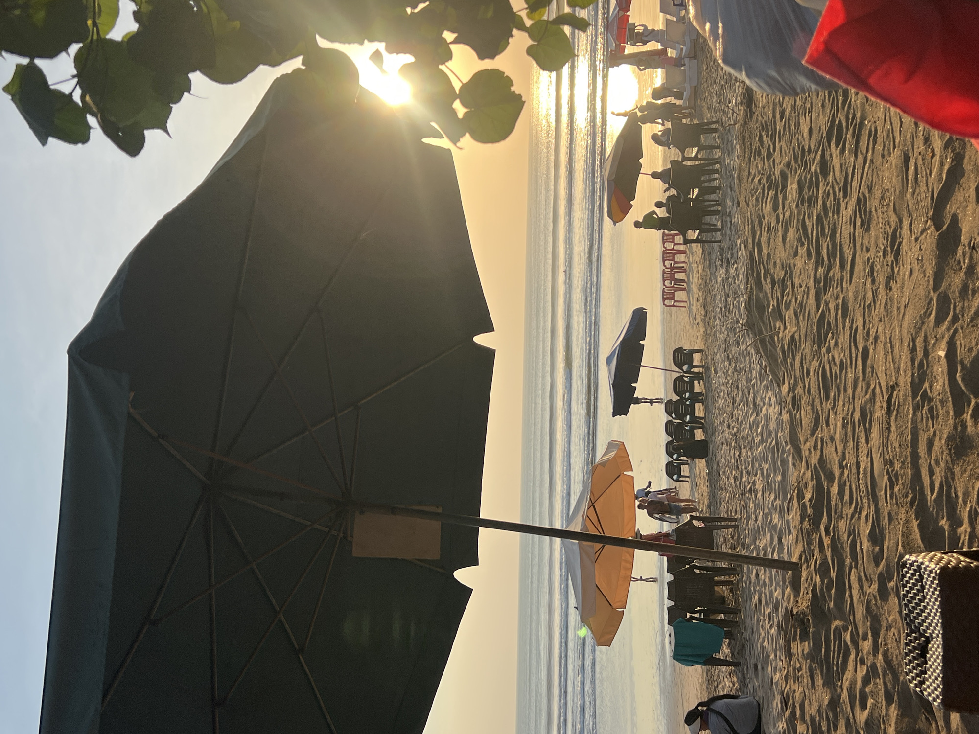 A sun-drenched beach scene unfolds in Kuta, Indonesia, as the golden hour casts a warm glow across the sand and sea. The foreground is dominated by the shadow of a large, dark green umbrella, its spokes partially visible. To its right, lush green leaves frame the radiant setting sun, which glints off the ocean's surface.
Scattered across the beach are numerous beach umbrellas, including a prominent orange and yellow one near the center, and several dark blue and red ones further back. Beneath these umbrellas, groups of people are visible, some seated at tables, others standing or strolling. Beach chairs are arranged in neat rows, awaiting visitors.
In the middle ground, the ocean stretches out, with gentle waves rolling towards the shore. A few figures can be seen in the water, enjoying the surf. The beach itself is composed of fine, light-brown sand, marked with the textured patterns of footprints and the tide.
The overall impression is one of a relaxed and idyllic beach setting at the end of the day, likely a popular spot for both locals and tourists to unwind and enjoy the natural beauty of Kuta.