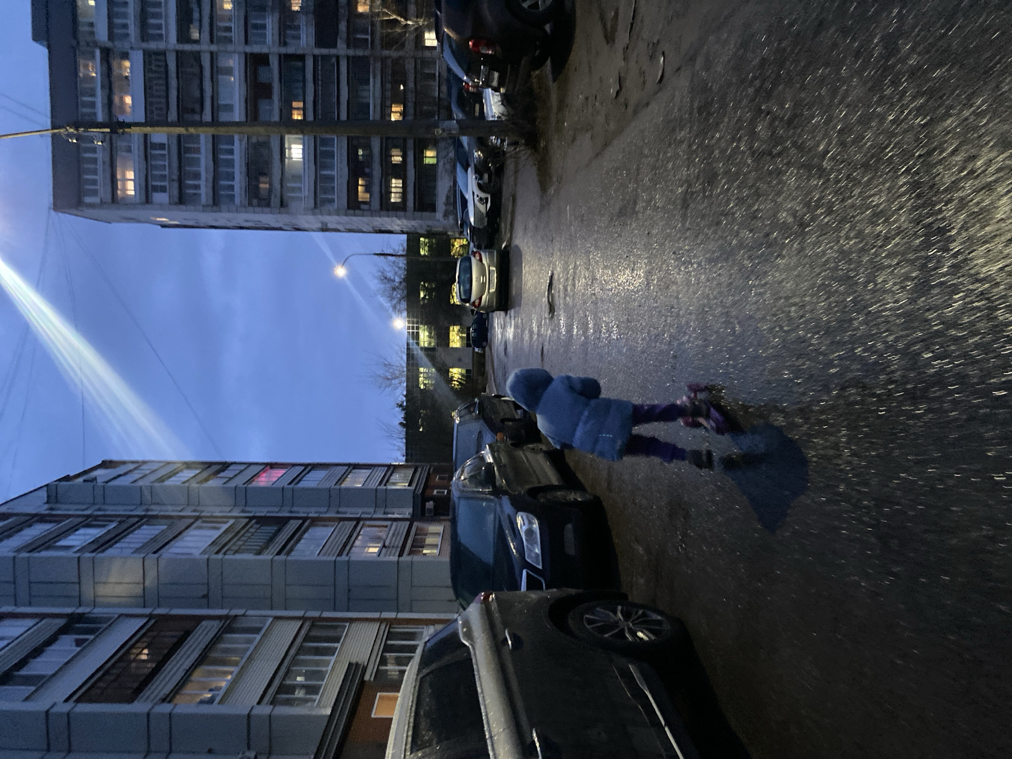 The image depicts an urban street scene in Chaikovsky, Russia, at dusk or early evening. The sky is a deep blue, indicative of twilight, with artificial lights beginning to dominate. The ground, likely an asphalt street or parking area, is wet and reflective, suggesting recent rain or snowmelt.
In the mid-ground, a child, dressed in a blue hooded jacket and dark pants, is in motion across the wet pavement. They appear to be riding a scooter or roller skates, with one foot lifted and a distinct shadow cast on the glistening surface behind them. A row of parked cars, varying in model and color, lines the street to the child's left.
The background is dominated by large, multi-story apartment buildings with numerous windows, many of which are illuminated, indicating residential activity. The architecture is utilitarian, characteristic of many Russian cities. Several streetlights are visible, glowing and casting reflections on the wet surfaces. A prominent, bright diagonal beam of light, possibly from a powerful streetlamp or vehicle headlight, cuts across the upper left of the frame. The overall atmosphere is quiet, capturing an everyday moment as night falls in a residential area. No discernible text is visible in the image.