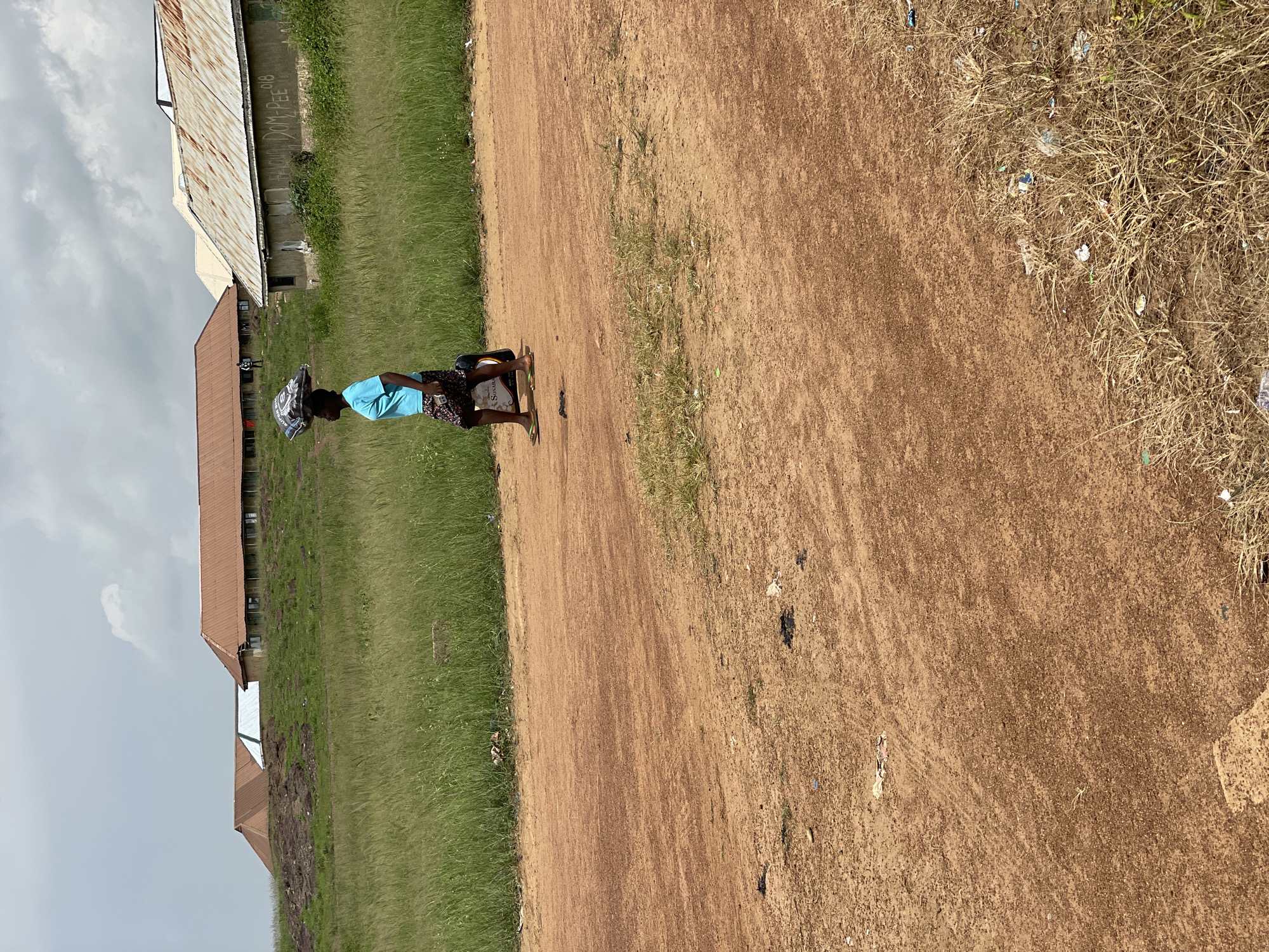 In Durumi, Nigeria, a young woman walks along a dirt road, carrying a large package on her head. She is wearing a blue t-shirt and a patterned skirt, and her feet are adorned with flip-flops. The road is a reddish-brown color, and it is bordered by a field of tall green grass. In the background, several buildings with corrugated metal roofs can be seen. The sky is cloudy, suggesting a humid and possibly rainy day. The overall scene depicts a moment of everyday life in a rural Nigerian setting.