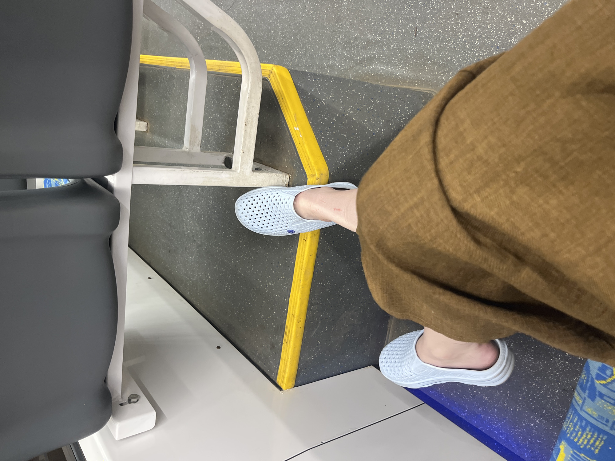 The image shows a person sitting on a bus or train, likely in Moscow, Russia, based on the style of seating and the flooring. The person is wearing brown trousers and light blue slip-on shoes. One foot is positioned near a yellow safety barrier, and the other is near the edge of a blue section. A gray bus or train seat is visible to the left of the frame. The flooring is a speckled gray pattern.
