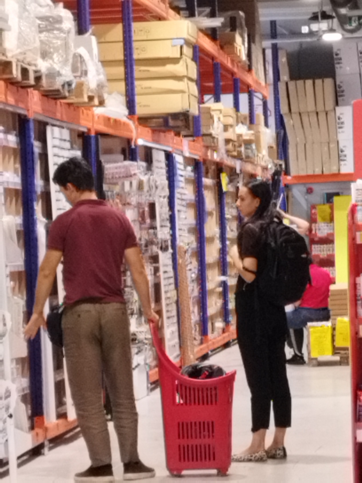 In a retail environment, possibly a hypermarket or warehouse store, two individuals are seen among shelves stocked with merchandise. A man in a maroon polo shirt and khaki pants stands facing away from the camera, his left hand resting on a red shopping basket. To his right, a woman with dark hair, wearing a black top and black pants, carries a black backpack and looks towards the left. Behind the woman, another person in a pink shirt and blue jeans is partially visible, seated on a stool. The shelves are filled with various items, including boxes, packaging, and possibly household goods. The flooring appears to be concrete or a similar industrial material. The lighting suggests an indoor commercial setting. The scene does not contain any specific indicators that place it in Bucharest, Romania.