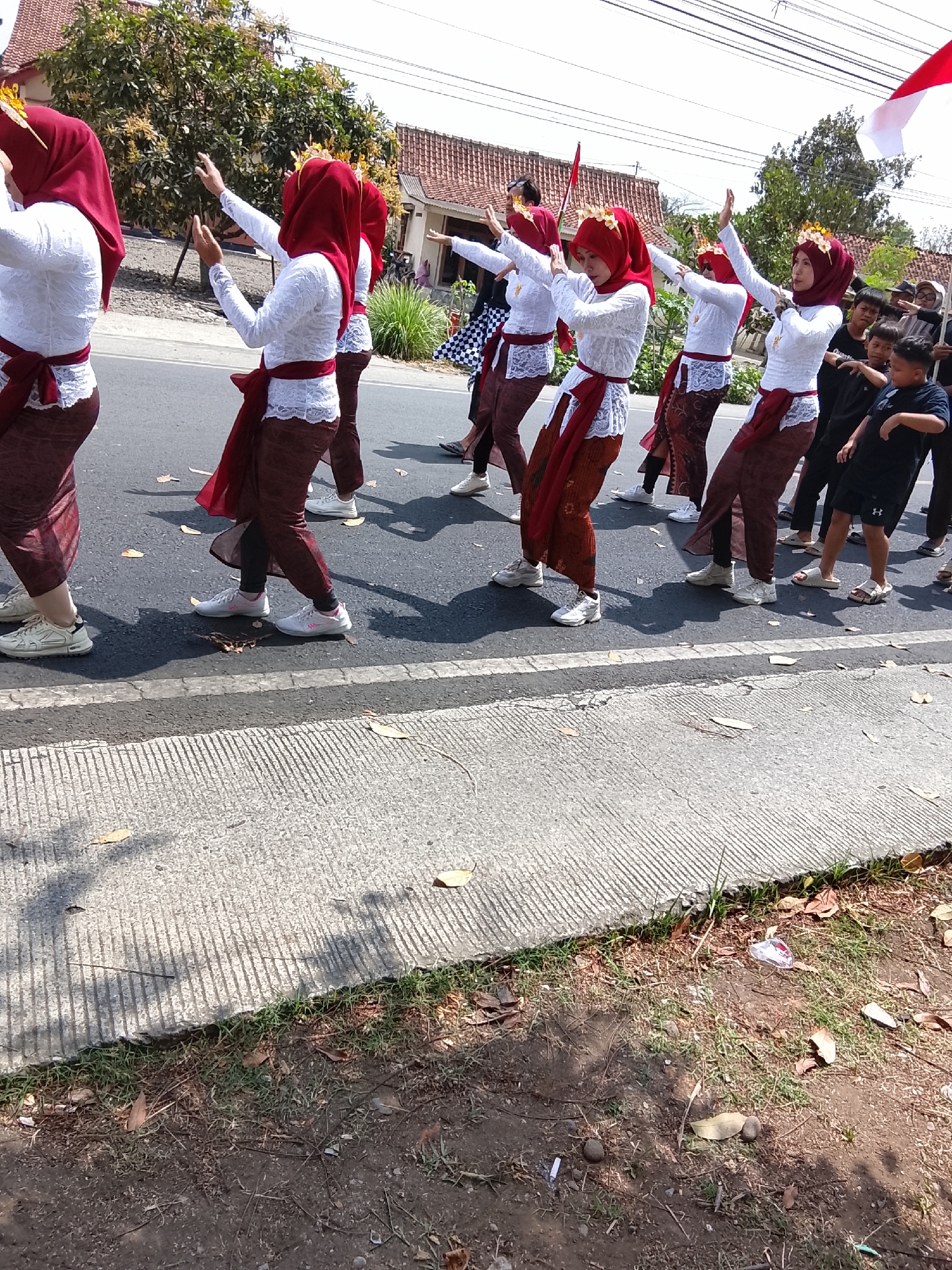 In Sampang, Indonesia, a group of young women in traditional attire participates in a parade. They are wearing white blouses with lace details, red hijabs, and patterned red skirts. Their outfits are accented with sashes tied at the waist and decorative headbands adorned with flowers. The women are performing a synchronized dance, with their arms raised in unison. Behind them, other participants and onlookers are visible, some holding Indonesian flags. The parade takes place on a paved road lined with trees and houses. The ground is dry and scattered with fallen leaves.