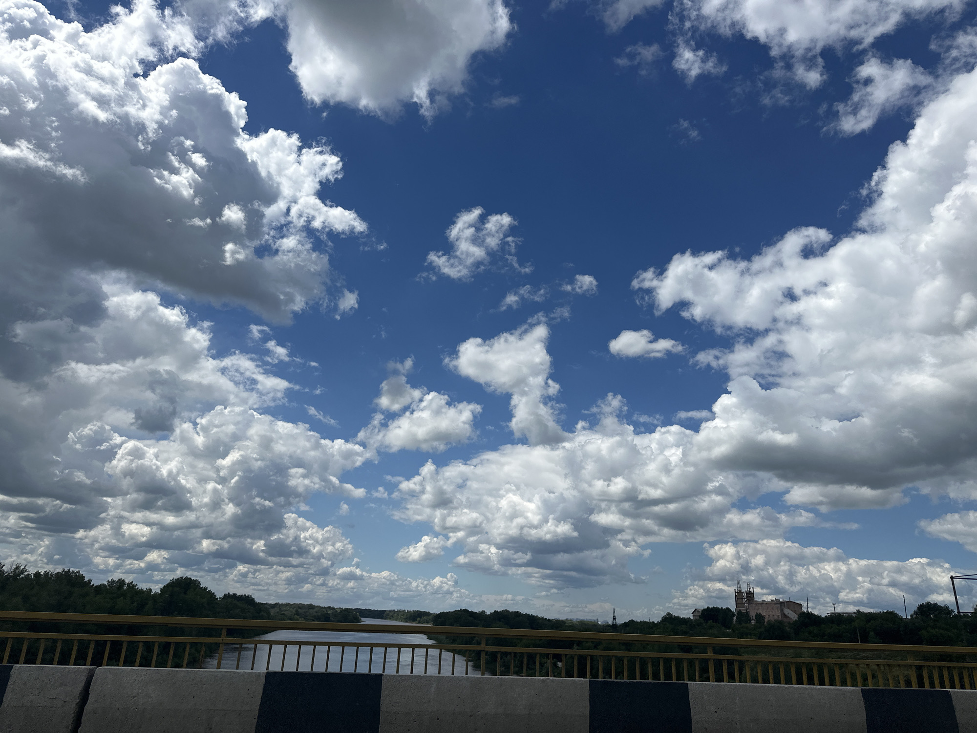 The image captures a view from a bridge overlooking a river, with a lush green tree line bordering the opposite bank. The sky is a vibrant blue, filled with dramatic, white cumulus clouds. In the distance, a cluster of buildings, possibly industrial, with prominent smokestacks, is visible against the horizon. The bridge itself is characterized by a yellow railing and a concrete barrier at the foreground. The overall scene suggests a bright, possibly overcast day in a semi-urban or rural environment. The location is identified as Alekseyevka, Russia.