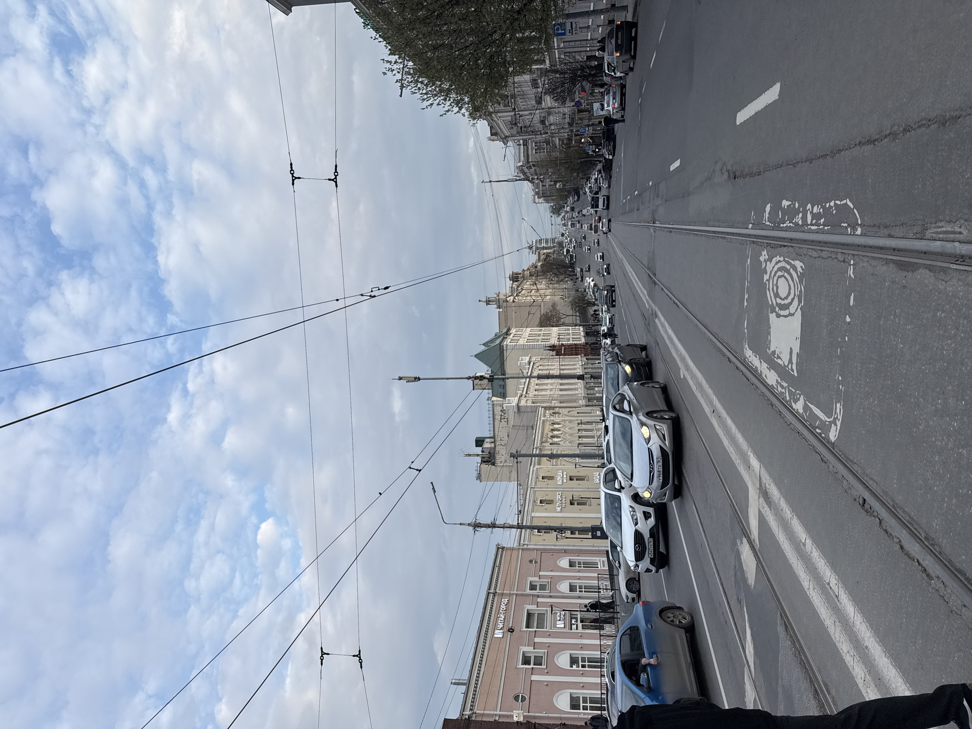 The image shows a street scene in Rostov-on-Don, Russia, with a long straight road stretching into the distance. The road features tram tracks running down the center, flanked by lanes of vehicular traffic. Cars are lined up in both directions. Overhead tram lines are visible, strung between poles along the street. The buildings lining the street are a mix of architectural styles, including older, ornate structures and more modern constructions. The sky is overcast with cloudy patches. There are few visible pedestrians.