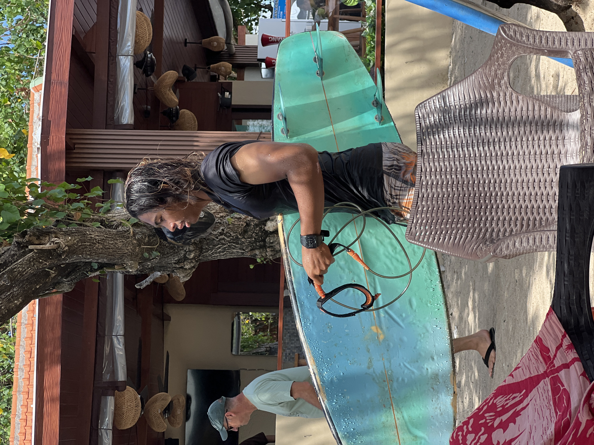 In Kuta, Indonesia, a young man with wet, dark hair, wearing a black short-sleeved shirt and patterned shorts, carries a light blue surfboard. He is walking away from the viewer, holding the leash of the surfboard with his right hand. A second individual, an older man wearing a green t-shirt and a green cap, walks past the young man. The scene is set on a sandy area, with a building and a tree in the background. The building features a reddish-brown roof and a textured wall, with straw-like hanging light fixtures. Beach chairs are visible in the foreground.