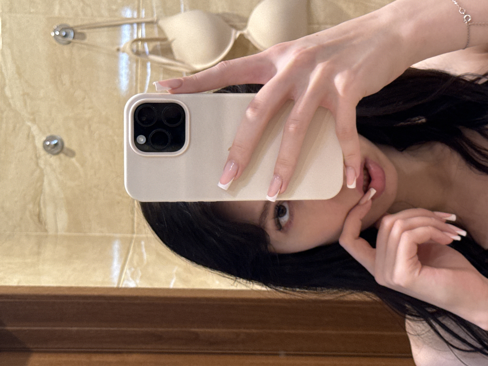 A young woman with dark hair takes a selfie in a bathroom. She is holding a light-colored smartphone in her right hand, obscuring the upper portion of her face. Her left hand is positioned near her mouth, with her index finger lightly touching her lips. She is wearing a light-colored top and a delicate silver bracelet on her left wrist. In the background, a beige bra is hanging on hooks against a tiled wall, and a wooden door frame is visible to the left. The lighting appears to be indoor, likely from a mirror or overhead light. The overall scene suggests a private, personal moment.