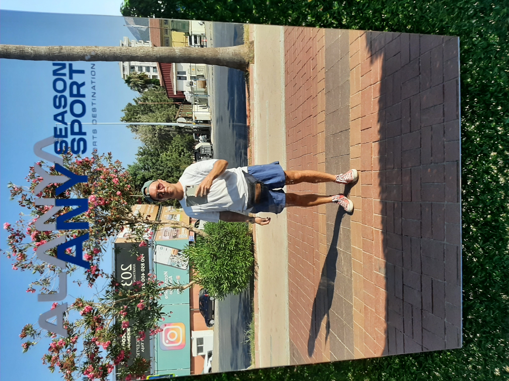 A man is taking a mirror selfie on a sunny day in Alanya, Turkey. He is wearing a white t-shirt, blue shorts, a fanny pack, sunglasses, and a baseball cap. He is standing on a brick sidewalk, with a street and buildings visible in the reflection of the mirror. The mirror is positioned in front of a large billboard with the words "ALANYA SEASON SPORT DESTINATION" in large letters. Trees with pink flowers are also visible in the reflection. The foreground of the image shows a patch of green grass.