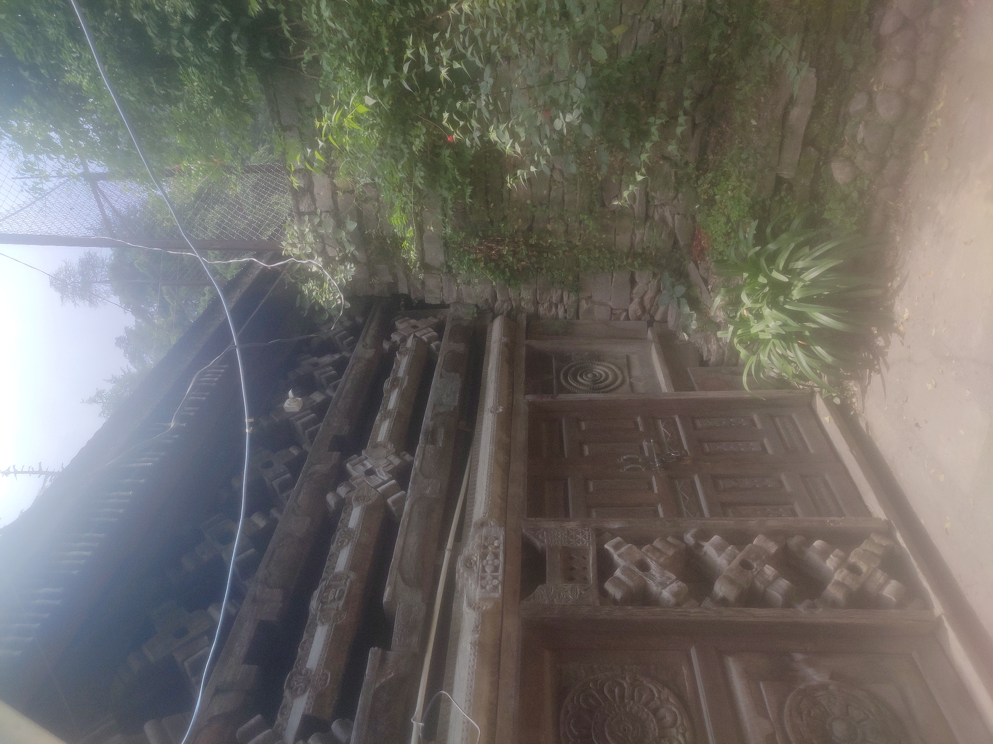 The image shows the intricately carved wooden facade of a traditional house in Naggar, India. The house features elaborate wooden pillars, decorative carvings on the doors, and a sloped roof. The exterior wall is made of stone and is partially covered by lush green foliage. A wire fence runs along the top of the stone wall, separating the property from the surrounding vegetation and trees. The scene is bathed in a soft, diffused light, possibly indicating an overcast day or early morning mist. The ground in front of the house is a dirt path.