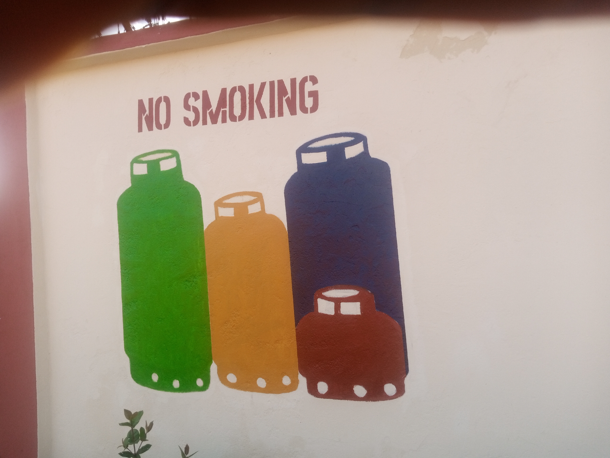 This media file features a painted mural on a light-colored wall. The mural depicts four stylized gas cylinders of varying sizes and colors: green, orange, dark blue, and brown. The cylinders have white accents on their tops, representing the valves or caps. At the base of the green and orange cylinders, there are white circular shapes, possibly representing feet or a design element.
Above the gas cylinders, the words "NO SMOKING" are stenciled in a bold, reddish-brown font. The letters are blocky and appear to be painted on with a stencil.
The wall itself shows some signs of wear, with patches of lighter discoloration visible, particularly towards the top of the image. The overall setting appears to be outdoors, as evidenced by the dappled light and a small plant with green leaves growing at the bottom left of the frame. There are no people visible in the media file. The time of day is indeterminate, and no weather conditions are evident. The context of the location is unknown, but the "NO SMOKING" sign and the depiction of gas cylinders suggest a place where safety regulations are important, possibly a facility or outdoor area. The image is taken at a slight angle, with a dark, out-of-focus area at the top that may be a person or object obstructing the view.