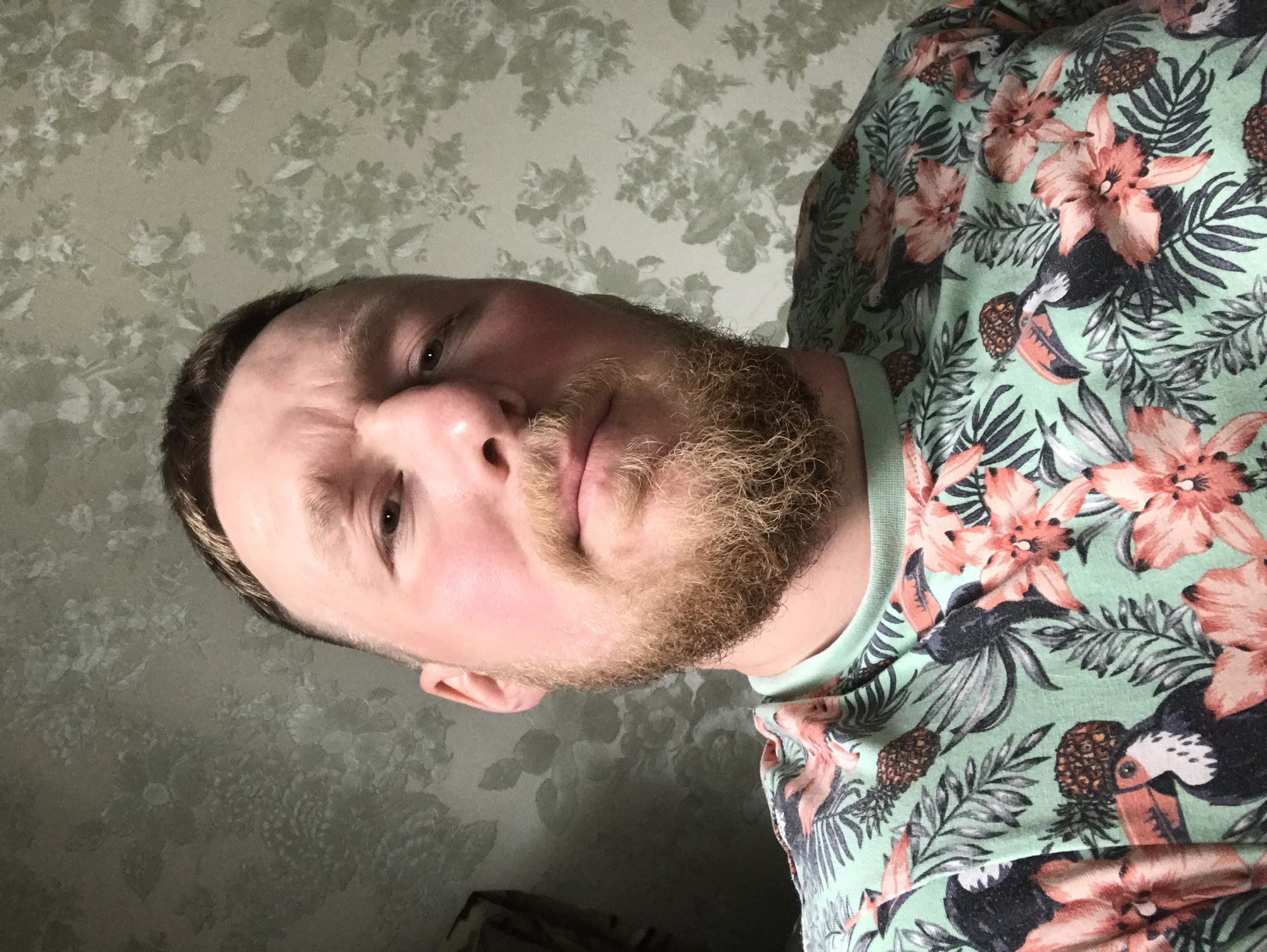 A man with a reddish complexion and a beard is the primary subject of the image. He is wearing a light green shirt patterned with tropical flowers, pineapples, and toucans. The background is a patterned wallpaper in shades of gray. The image appears to be a close-up or selfie, taken indoors. The location is unknown.