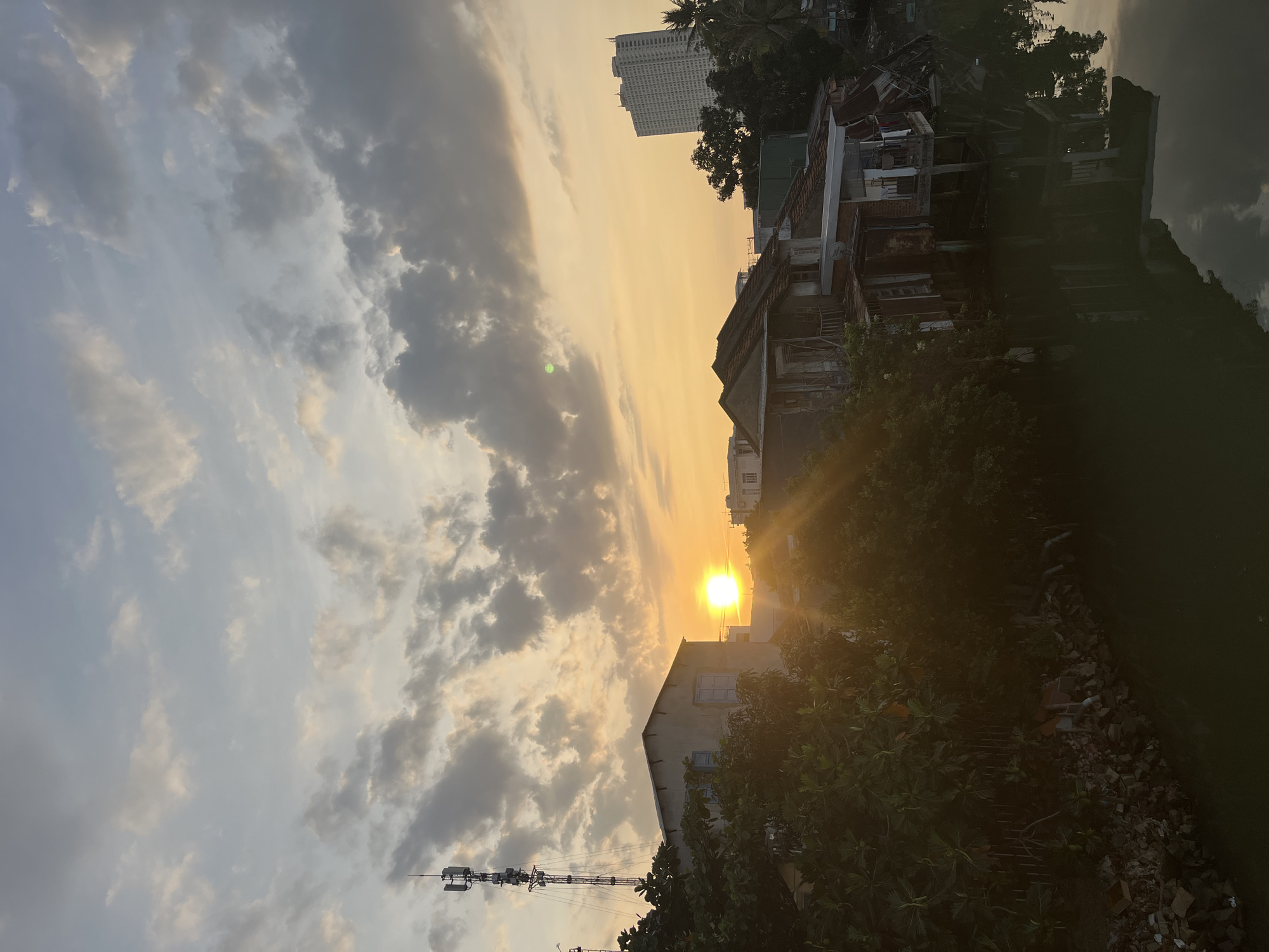 The image depicts a sunset in Nha Trang Ward, Vietnam. A bright sun is positioned between buildings and trees, casting a golden hue across the sky and the clouds. The architecture varies, with some older, dilapidated buildings alongside a modern high-rise structure. The foreground is dominated by vegetation, some of which obscures the view. A communications tower is visible in the lower left corner. The image is rotated sideways.
