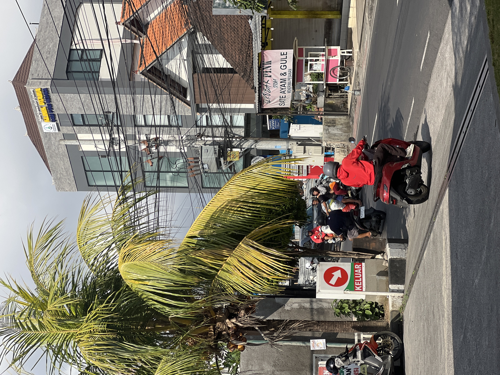 This image captures a street scene in Denpasar, Indonesia, under a bright, partly cloudy sky. In the foreground, a person wearing a red hoodie and dark pants rides a red scooter, facing away from the camera and moving rightward. The scooter has a license plate reading "DK 3330 JW". Several other scooters and their riders are visible behind this central figure, some stationary and some moving. To the left, a mature palm tree with coconuts is prominent, its fronds casting shadows on the pavement.
Behind the palm tree, a sign with a red arrow pointing right and the word "KELUAR" (Indonesian for "EXIT") is mounted on a concrete post. Further back, a modern, multi-story building with grey brick siding and large windows stands to the right of a smaller building with a red-tiled roof. This larger building appears to be a clinic, as indicated by the signage on its top that reads "KLINIK UTAMA" and "PAN PURI MEDIKA". Numerous electrical wires crisscross the scene, strung between poles and buildings.
To the right of the clinic, a small food stall with a pink and white cart is set up. A banner above it displays "SEDAP PINK SATE AYAM & GULE MENERIMA PESANAN" (Indonesian for "Delicious Pink Chicken Satay & Gule - Orders Accepted"). The street is paved with asphalt, marked with white lines, and bordered by a raised concrete sidewalk. The overall atmosphere suggests a typical urban day in Denpasar, with traffic, commerce, and distinct Balinese architectural elements.