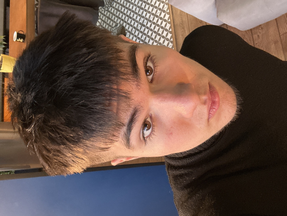 The image displays a close-up, high-angle portrait of a young male, centered in the frame. The subject has light skin, brown eyes, and short, dark brown hair with a faded cut on the sides. He wears a black turtleneck and exhibits faint stubble on his chin and upper lip. His gaze is directed straight at the camera.
The background depicts an indoor environment. To the subject's immediate left, a portion of a vertical blue surface, possibly a wall or screen, is visible, illuminated by a warm light source. Above and to the subject's right, a wooden surface, likely a table or shelf, holds a small yellow plant pot containing green foliage and a black mug with a white design. A dark brown fabric object is partially visible behind the mug. The floor below consists of white and black geometric patterned tiles, transitioning to light-colored wooden planks. A section of light-colored, possibly fabric, furniture is visible on the far lower right. The specific location within Kyiv, Ukraine, cannot be determined from the visual information provided.