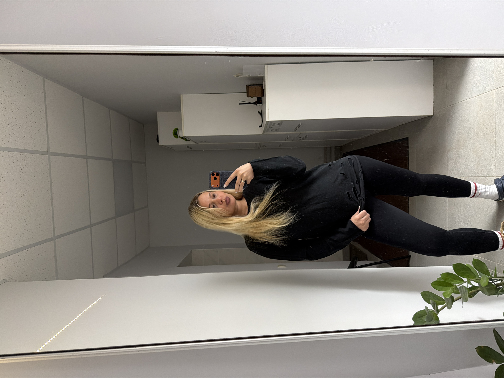 A woman stands in front of a mirror taking a selfie. She is wearing a black hoodie and black leggings with white and red striped socks. She has long blonde hair. The background shows a bathroom or kitchen area with white cabinets and tiled floors. The specific location in Sarapul, Russia cannot be determined from the image.