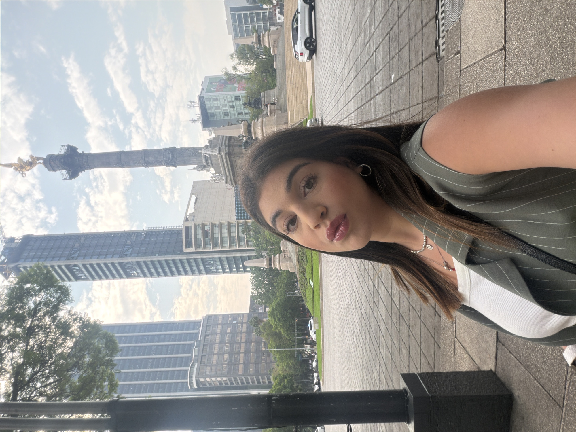 A selfie shows a young woman with long brown hair wearing a green pinstripe vest, a white top, and jewelry. The background features a cityscape that includes the Ángel de la Independencia monument in Mexico City, Mexico. Modern high-rise buildings and other structures surround the monument under a cloudy sky. The ground appears to be paved, likely a pedestrian area or walkway. A white car is visible.