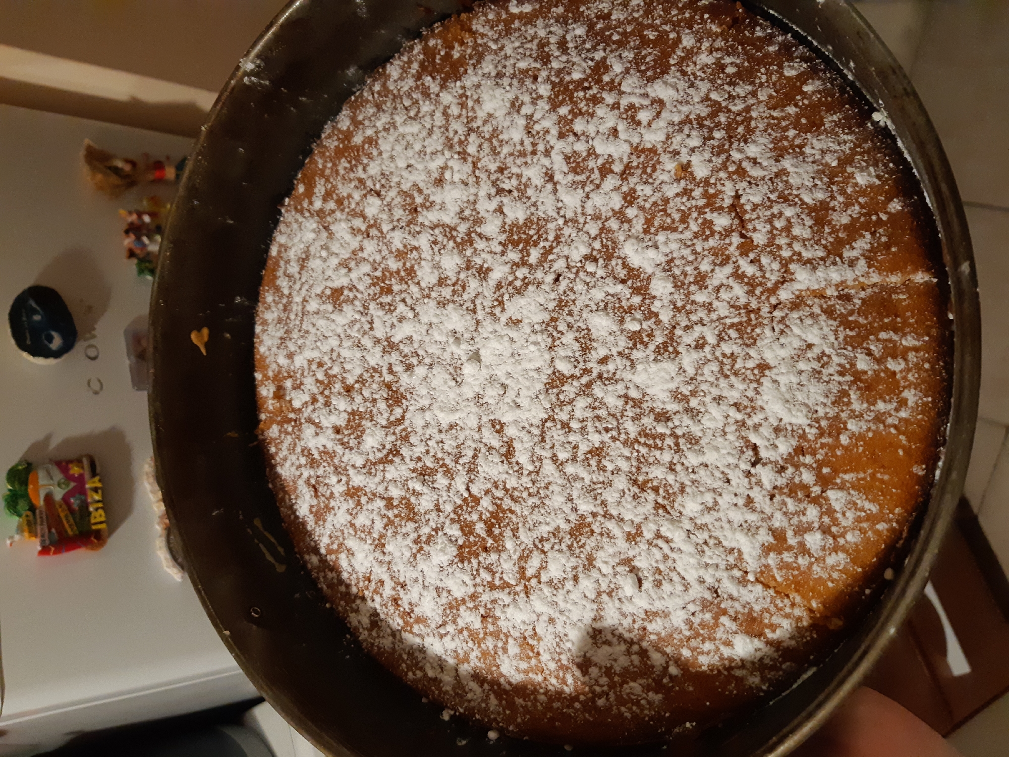 The image shows a round cake in a metal baking pan. The cake is covered in a layer of powdered sugar. The pan appears to be sitting on a white surface. In the background, there are several refrigerator magnets, including one with the word "IBIZA". The image does not show any information that would suggest it was taken in Plovdiv, Bulgaria.