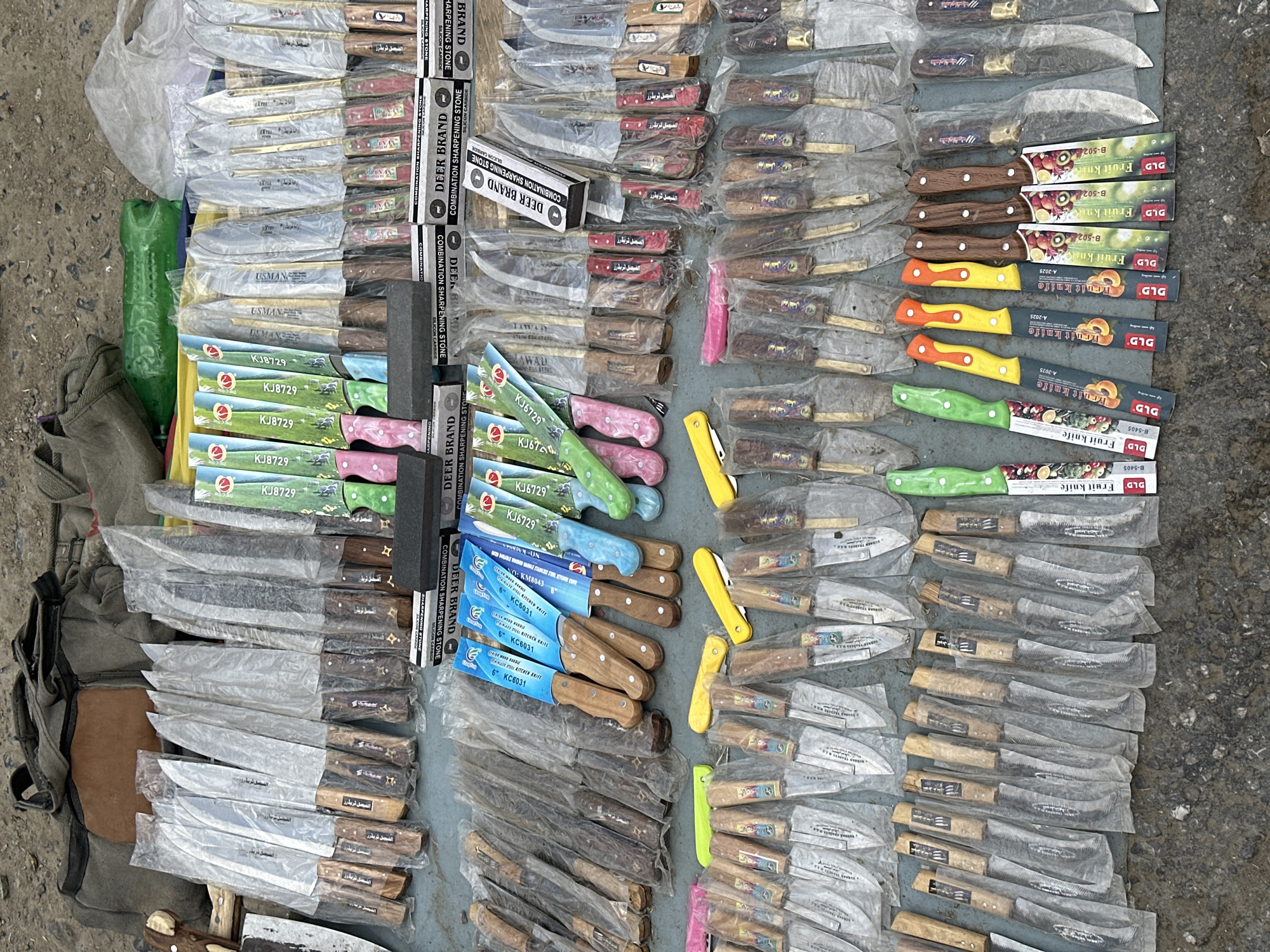 The image shows a collection of knives and sharpening stones laid out on a surface, likely for sale. Many of the knives are individually wrapped in clear plastic. There are several different styles of knives including small folding knives, fruit knives, and larger blades. Some of the knives have colored handles (pink, green, yellow, orange, and blue). There are multiple boxes of "Deer Brand" combination sharpening stones. Some knives have packaging labeled in Arabic. The location is suggested by the packaging of some knives which indicates that they are from the city of Quetta City Tehsil, Pakistan.