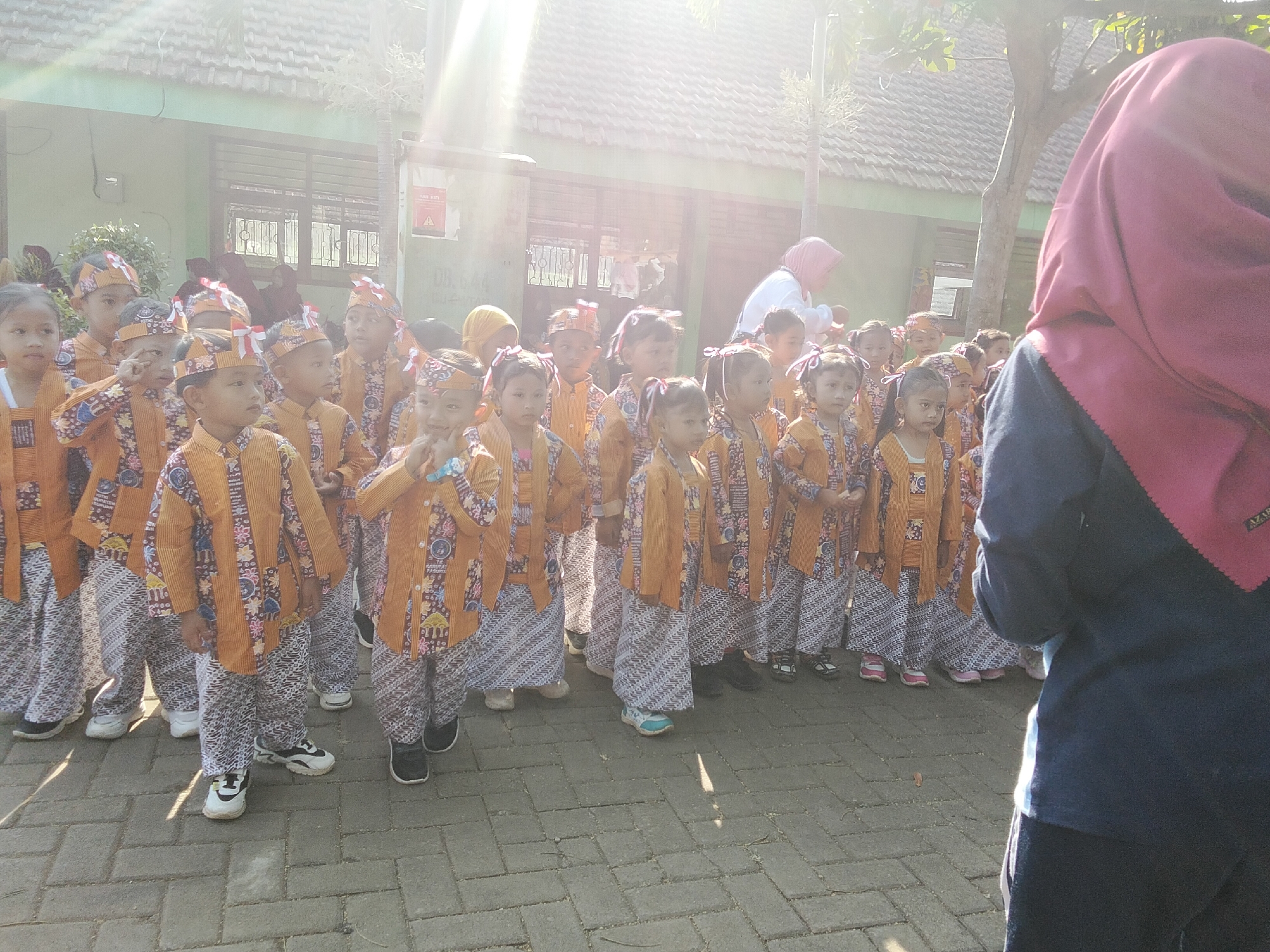 In Puspo, Indonesia, a group of young children, likely students, stand in formation outdoors. They are dressed in traditional attire, featuring patterned orange tops and pants, with some wearing matching headbands. The children are arranged in rows, facing forward, and appear to be engaged in an activity or waiting for instructions. Behind them, a building with a tiled roof and windows is visible, suggesting an educational institution. Sunlight streams down, casting shadows and creating lens flares in the image. A woman wearing a pink hijab is partially visible in the background, attending to the children. The ground is paved with brick-like material. The scene is likely a school event or cultural celebration.