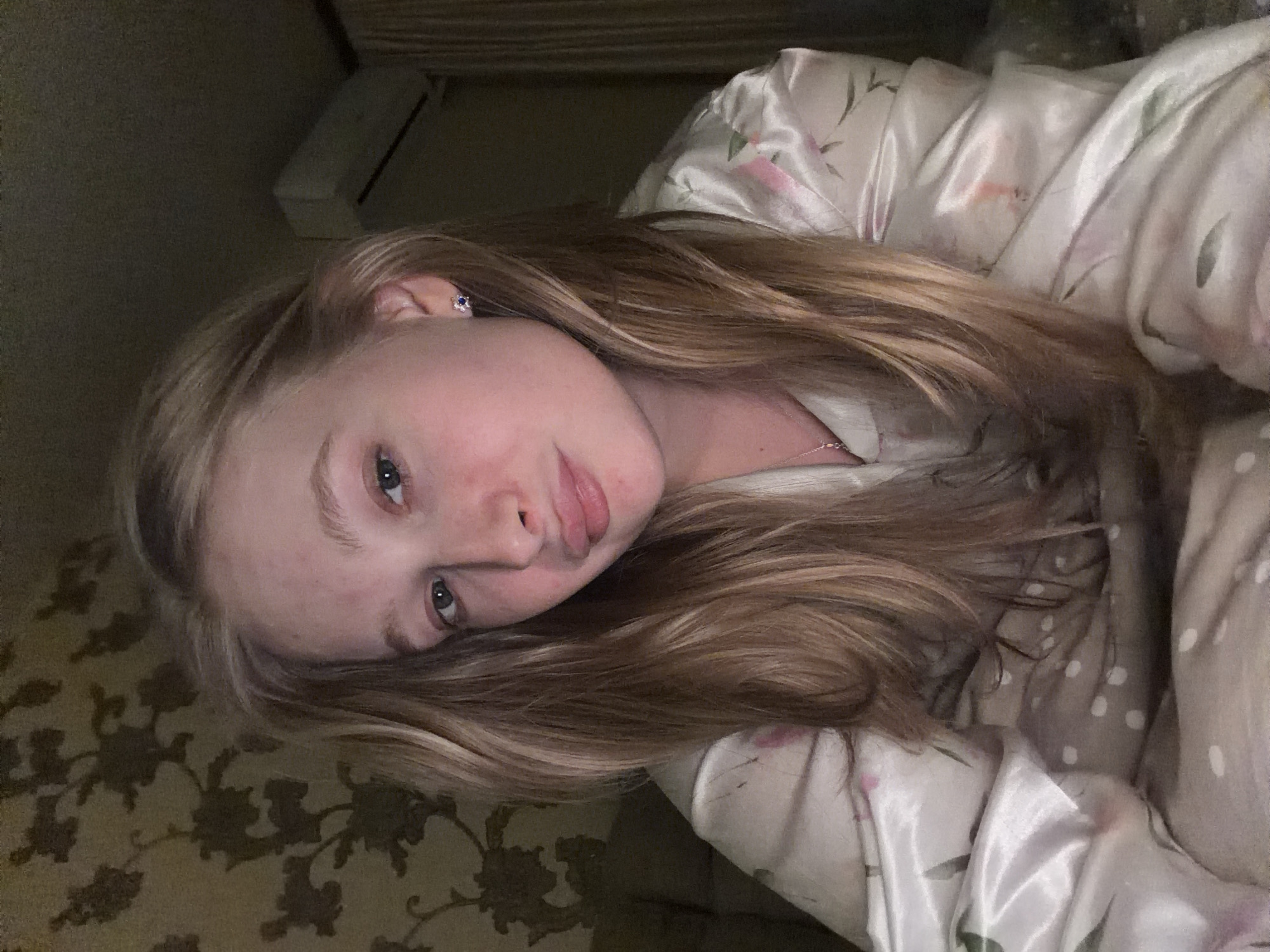 A pale-skinned individual with blonde hair is lying down on a patterned bed, wearing light-colored floral-patterned pajamas. The individual is facing the camera. A piece of furniture, possibly an air conditioner, is visible in the background. The location is Penza, Russia.
