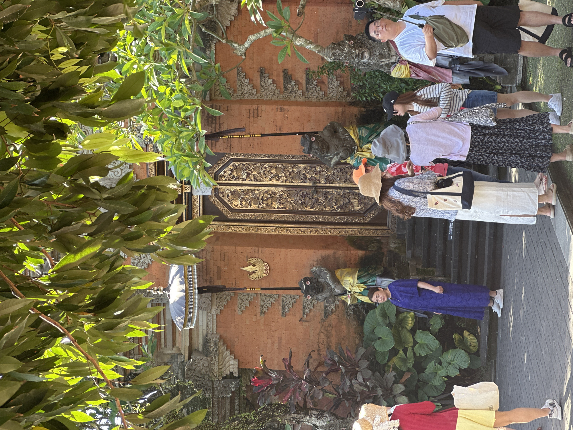 In the city of Ubud District, Indonesia, a group of tourists are gathered in front of a traditional Balinese temple entrance. The temple features intricate carvings and ornate golden doors. Two stone statues, resembling mythical creatures, guard the entrance. Overhanging trees with lush green leaves cast dappled sunlight on the scene. Several people are dressed in light, casual clothing, some wearing hats and carrying bags, suggesting a warm, sunny day. One woman in a vibrant blue dress stands out, looking towards the temple. Another woman, wearing a yellow skirt and red top, is partially visible on the left. The ground is paved with stone, and the overall atmosphere is one of cultural exploration and tourism.