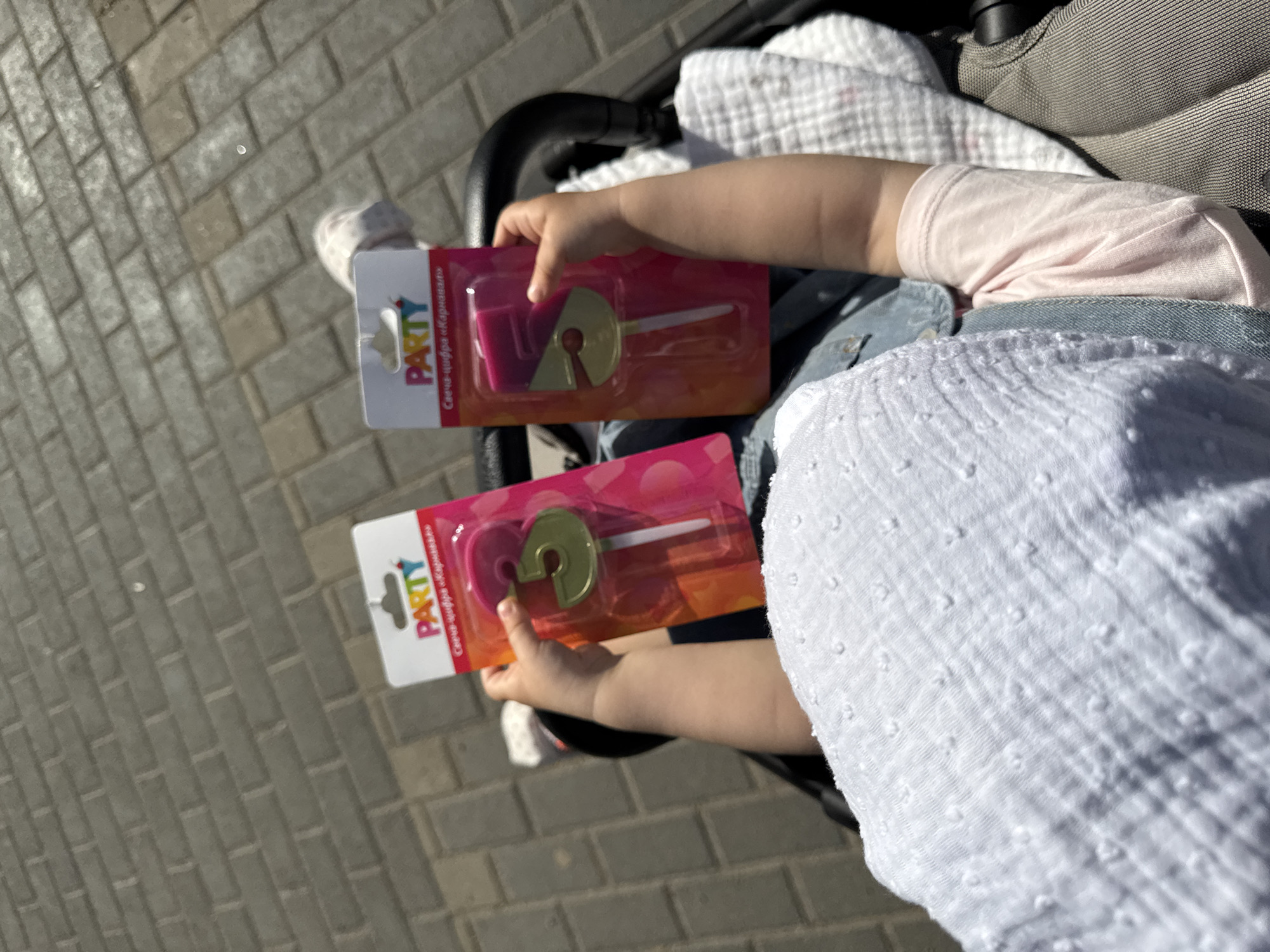A high-angle, medium shot depicts a baby holding two packages of birthday candles while sitting in a stroller. The packages are pink, white, and orange, with the word "PARTY" printed in bold, colorful letters. Each package contains a number-shaped candle, one a "7" and one a "3". The baby wears a white, textured dress and a pink shirt. The stroller appears to be a dark grey or black color. The background is composed of grey brick pavement, indicating an outdoor setting, possibly a street or sidewalk.