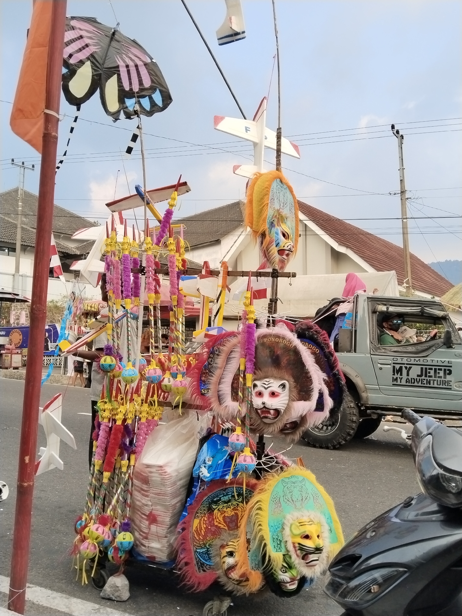 In the city of Nglebak, Indonesia, an outdoor scene unfolds with a street-side vendor's cart overflowing with colorful, traditional Indonesian masks and toy airplanes. The cart is laden with various items, including two distinct large masks with yellow and orange fur and detailed facial features, one with a tiger motif. Smaller, hanging ornaments with spherical, multi-colored elements adorn the cart. Several toy airplanes, predominantly white with red accents, are displayed on poles. A large, black butterfly kite with pink and yellow markings is suspended above the cart. A light gray SUV, branded "MY JEEP MY ADVENTURE," is parked nearby, with a person inside wearing sunglasses and holding a white object. A black motorcycle is partially visible in the foreground. The backdrop consists of residential buildings with red tile roofs, utility poles with overhead wires, and a cloudy sky. The street is paved, and a white line marks the edge of the road.