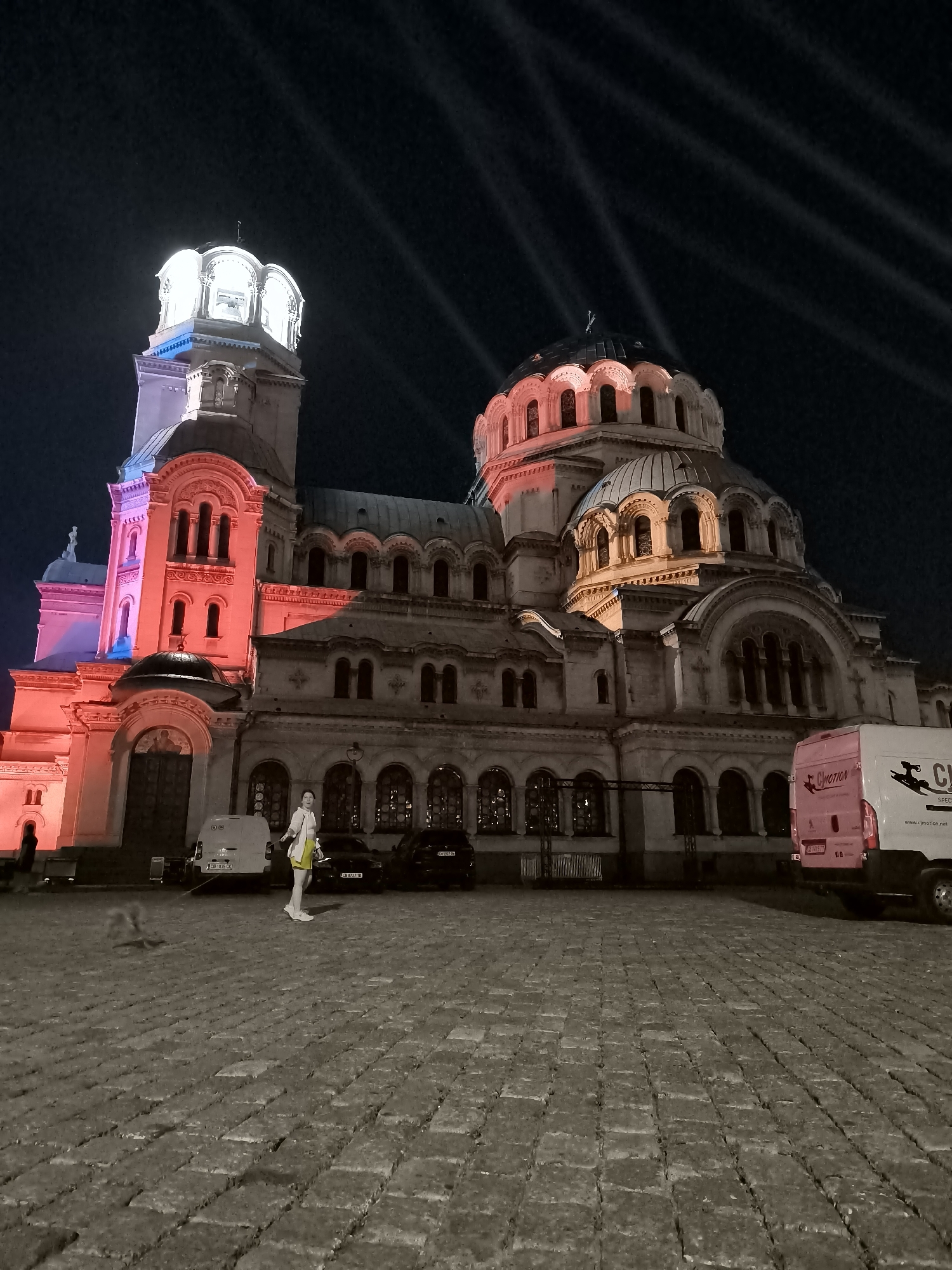 The image shows the exterior of the Alexander Nevsky Cathedral in Sofia, Bulgaria, at night. The cathedral's facade is illuminated with red and blue lights, highlighting its architectural features. A woman is standing in the foreground, facing away from the camera and to her left. She is wearing a white shirt, yellow shorts, and white sneakers, and carrying a yellow bag. Two cars and a white van are parked in front of the cathedral. The ground is paved with cobblestones. Light beams are visible in the dark sky above the cathedral.