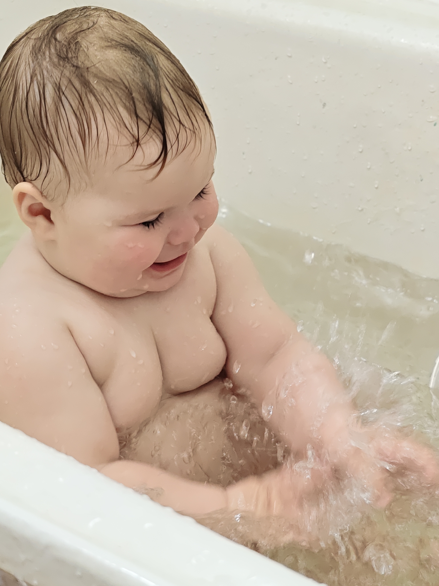 A baby is sitting in a bathtub filled with water, splashing with both hands. The baby has wet hair and appears to be smiling while looking down at the water. The bathtub is white and the surrounding environment is not visible. The location in Chaikovsky, Russia is not discernible from the image.