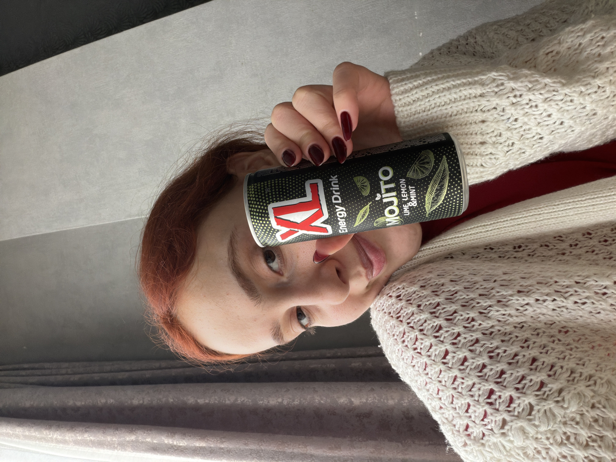 A woman with red hair and dark red nail polish is holding a black and green "XXL Energy Drink Mojito Lime, Lemon & Mint" can. She is wearing a white, knitted sweater over a red shirt. The background includes a gray curtain and a black and white wall. No information is present indicating the location as Tbilisi, Georgia.