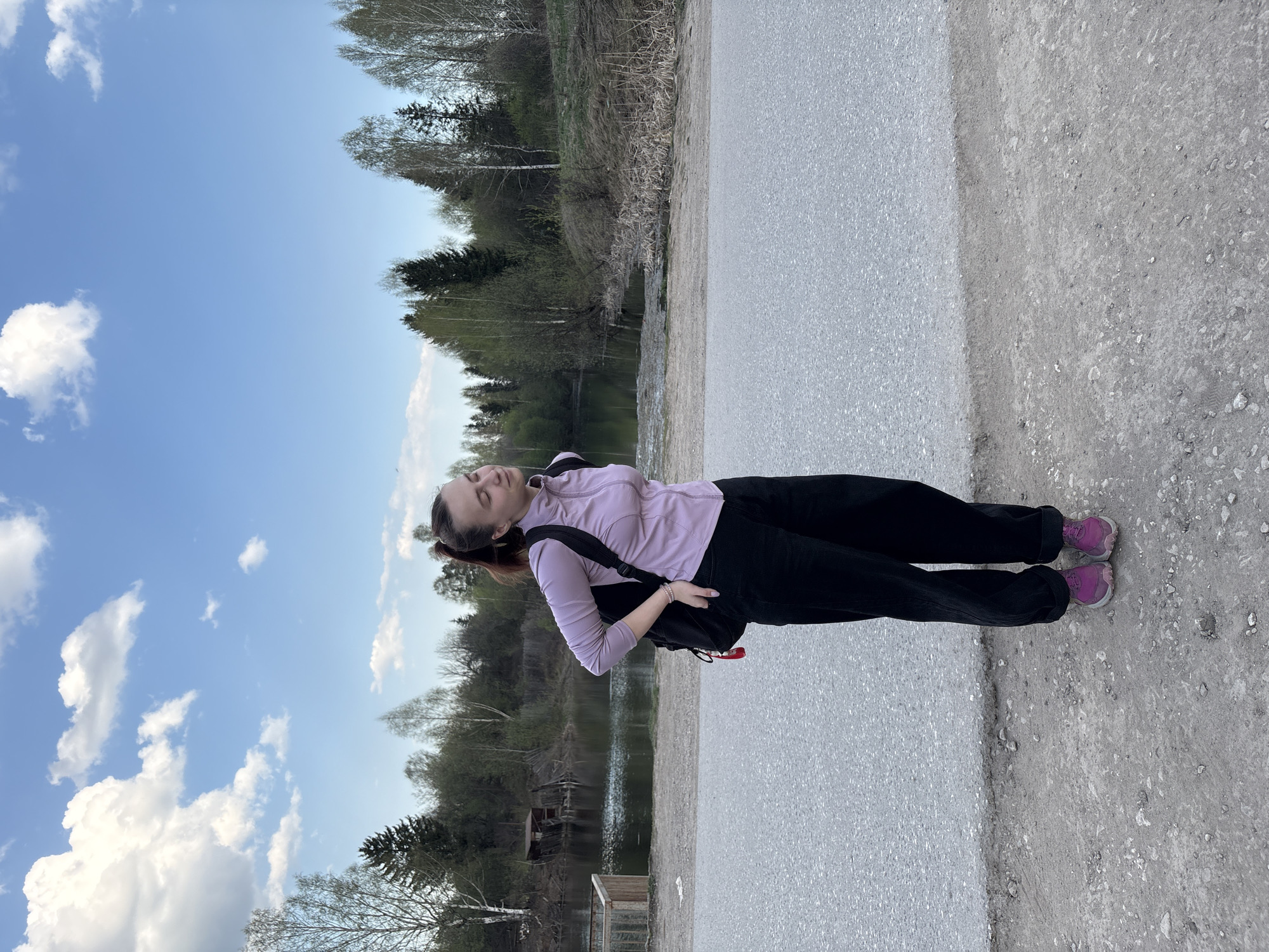 A woman stands on a paved path adjacent to a body of water in Perm, Russia. She is wearing a light purple long-sleeved shirt, black pants, and purple sneakers. A black backpack is slung over her shoulder. The path is bordered by a dirt and gravel area. Beyond the water, there is a wooded area with various trees. The sky is blue with scattered white clouds. In the background, near the water's edge, is a small wooden structure.