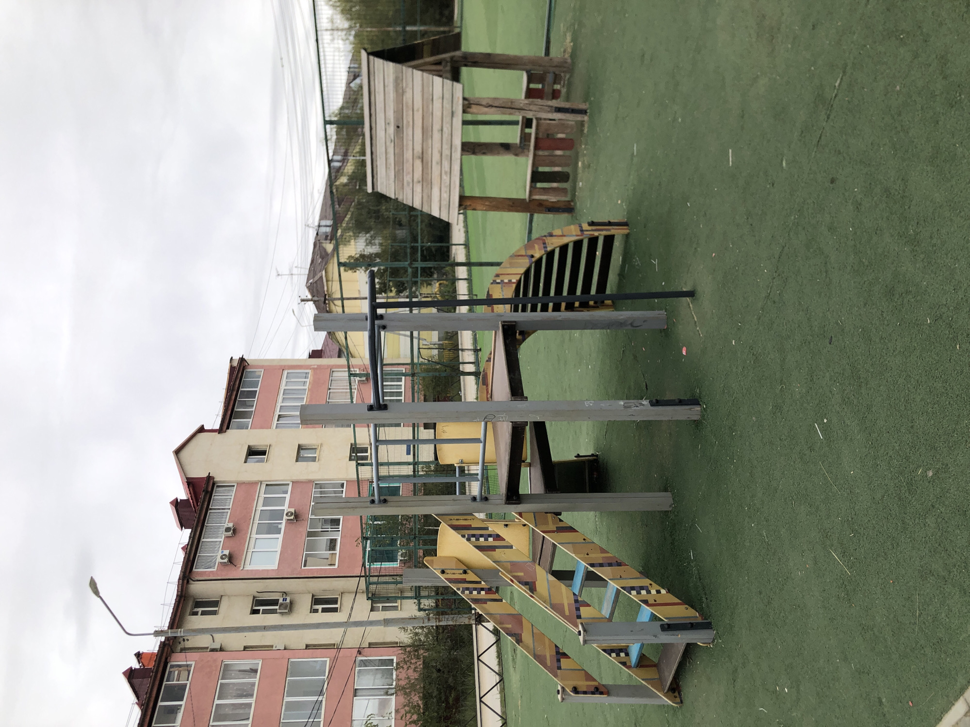 An outdoor playground, likely in Yakutsk, Russia, features a multi-level wooden and metal play structure with yellow and brown patterned stairs and a slide. To the right, a small, rustic wooden playhouse with a pitched roof stands on a green artificial turf surface. In the background, a pale pink and beige apartment building with many windows and air conditioning units is visible, along with a chain-link fence and some trees. The sky is overcast, suggesting a cloudy day. No people are present.