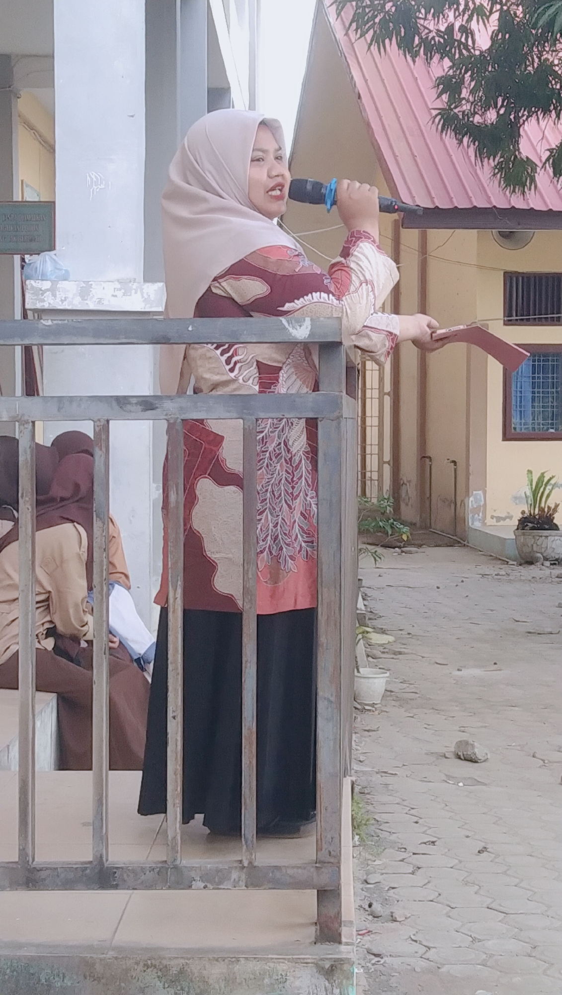 A woman wearing a beige hijab and a maroon and beige patterned blouse stands on a raised platform behind a metal railing, holding a microphone in her right hand and a small, reddish-brown rectangular object in her left. Her mouth is open, as if speaking into the microphone. Below her, to the left, two other individuals wearing hijabs and light brown uniforms are seated, partially obscured by the railing. The background shows a building with a pink roof and light yellow walls, a window with a blue grid, and some potted plants. The ground is paved with textured grey tiles and sand. The scene appears to be outdoors, possibly in a schoolyard or courtyard in Rhieng Blang, Indonesia.