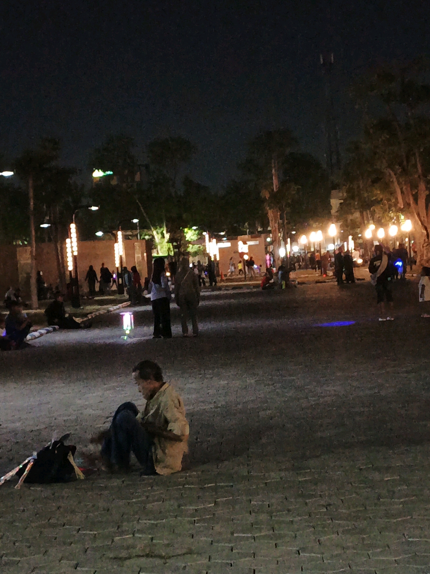 An outdoor night scene in Sidoarjo, Indonesia shows a group of people gathered in a public space that appears to be a park or plaza. The ground is paved with interlocking stones. Several tall, thin streetlights with vertical rows of illuminated bulbs line the pathway, casting a warm glow. In the foreground, a man is seated on the ground, facing away from the camera, with a dark backpack and some colorful, possibly reflective, objects beside him. In the middle ground, several figures are walking and standing, some illuminated by the streetlights. Further in the background, more people are visible, and the scene is lit by a variety of lights, including what appear to be decorative lights on buildings or structures. The overall atmosphere is one of a bustling evening gathering.
