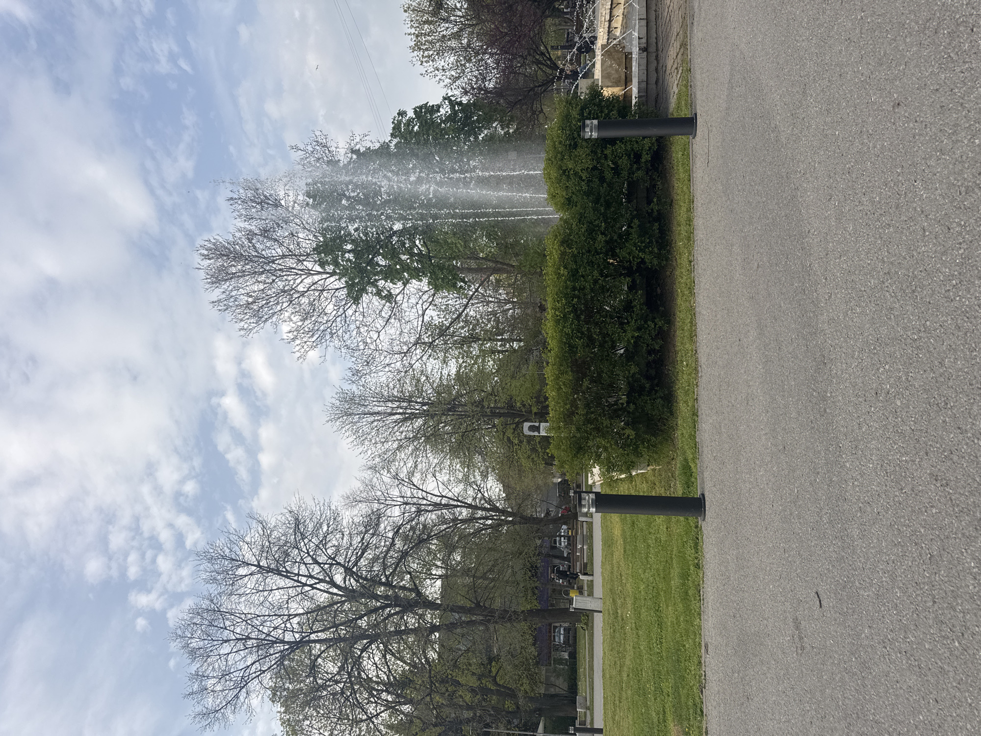 The image depicts a street scene in Montana, Bulgaria. A grey asphalt road occupies the right side of the frame, bordered by a strip of green grass and lined with short, black, cylindrical light posts. To the left of the road, there is a park area with trees in varying stages of foliage. A water fountain is active, spraying water high into the air near the treetops. Beyond the fountain, glimpses of buildings, tombstones, and possibly people are visible in the background. The sky is partially cloudy with a mix of blue and white. Overhead power lines are present in the sky.