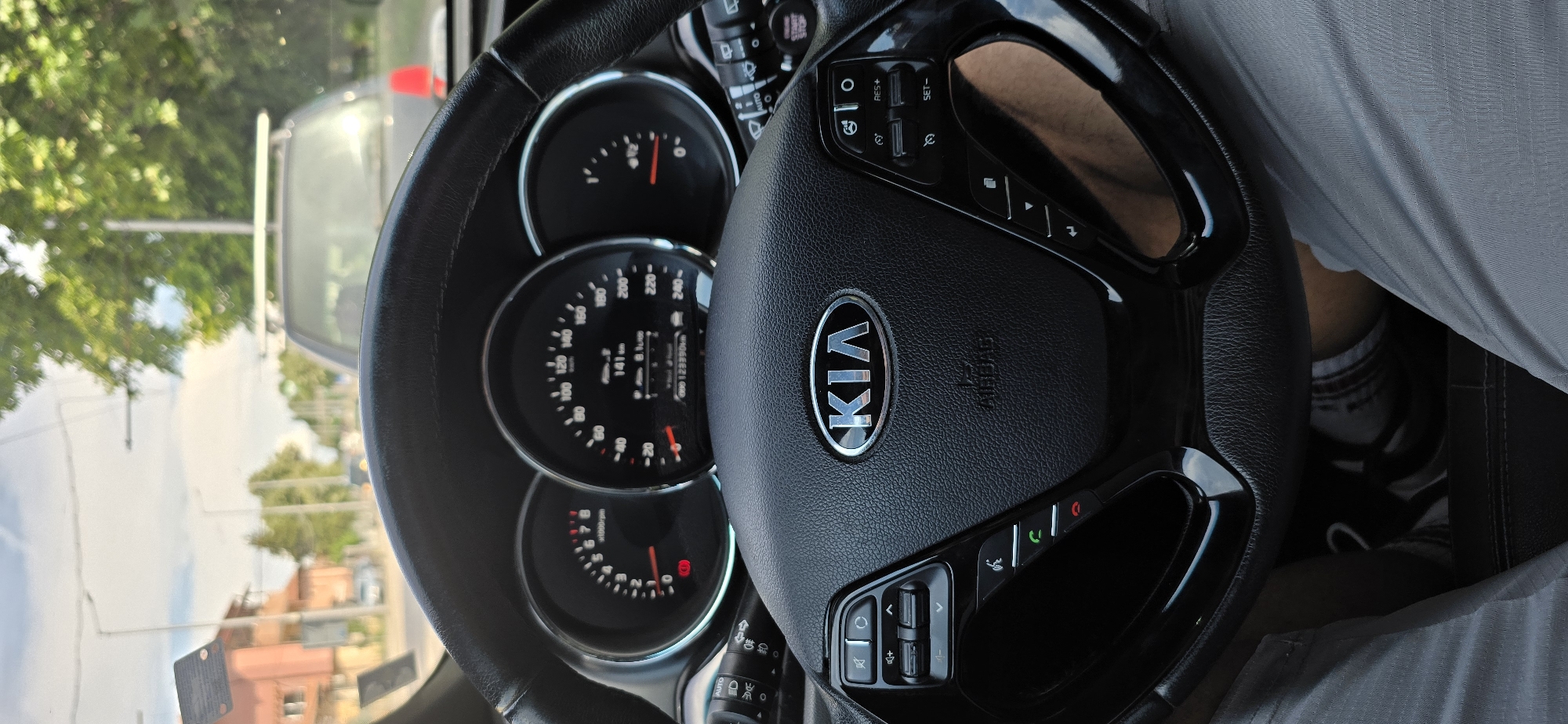 The image shows the interior of a Kia vehicle, looking forward through the windshield. The steering wheel is in the foreground, with the Kia logo prominently displayed. The dashboard features two circular gauges, one showing RPM and the other speed, with a digital display showing 141 km and 8.1 L/100km. The odometer reads 00123390km. The driver's legs are visible below the steering wheel, wearing light-colored shorts and athletic shoes. The exterior view through the windshield shows a street scene with buildings and trees. The vehicle appears to be in traffic or stopped.