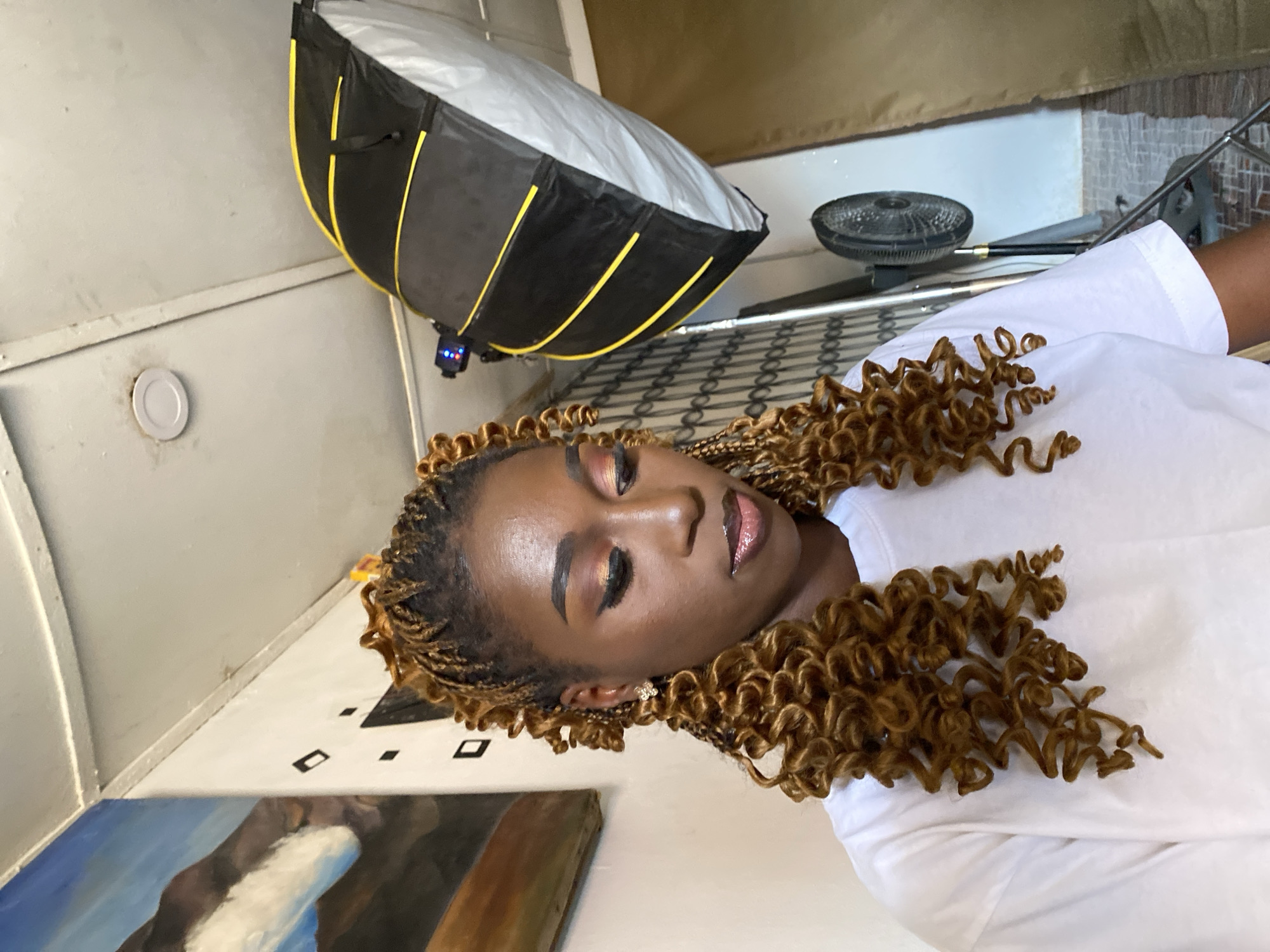 A close-up, portrait-oriented photograph depicts a young Black woman posing indoors, likely in Vom, Nigeria. The woman, positioned centrally, is lying on her back, looking towards the top left of the frame (if viewed upright). She wears a simple white t-shirt and has elaborate, light brown, curly and braided hair that frames her face and spills onto her shoulder. Her makeup is prominent, featuring shimmering eyeshadow in warm tones (orange/gold), defined eyebrows, and glossy lips. She has a serene, slightly alluring expression with her eyes partially closed. A small, decorative earring is visible in her left ear.
The background suggests a makeshift photography setup. To her right, a large, black and yellow photography softbox (octabox) with a white diffuser is suspended, providing key lighting. Below the softbox, a small, dark fan is visible. Further back, a metal structure resembling a bed frame or shelving unit is present. The walls are light-colored, and a section behind her head features a decorative pattern of black squares and diamonds on a white surface. A partially visible painting with blue and brown tones rests on the floor or a lower surface to her left. The overall lighting is soft, likely from the artificial source. No visible text is present in the image.