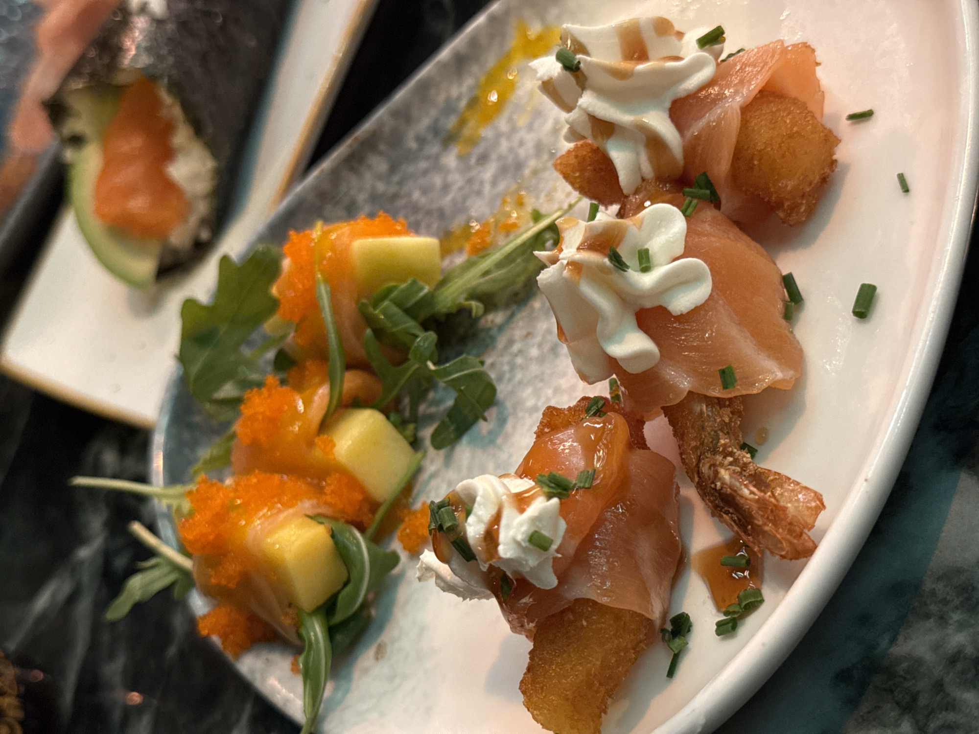 The image depicts a close-up view of multiple food items arranged on plates within a dining establishment in Barcelona, Spain. In the foreground, an oval white plate displays three individual portions. Each portion consists of a golden-brown, fried rectangular piece, potentially bread or potato, wrapped in a thin, light orange slice of cured fish. These are each topped with a piped swirl of white cream, drizzled with a translucent amber-colored sauce, and garnished with finely chopped green chives. One of these preparations also incorporates a fried shrimp tail. Adjacent to this, on a greyish-blue, irregularly shaped plate, are several yellow fruit cubes, identified as mango, interspersed with green arugula leaves. These elements are liberally covered with numerous bright orange fish roe particles. A similar amber sauce is present on this plate. In the out-of-focus background, a portion of a white rectangular plate reveals a sushi hand roll, partially wrapped in dark nori seaweed, containing white contents, orange fish, and a slice of green avocado. The overall scene indicates an indoor setting, possibly a restaurant, with artificial lighting focused on the food. No individuals or specific actions are visible.
