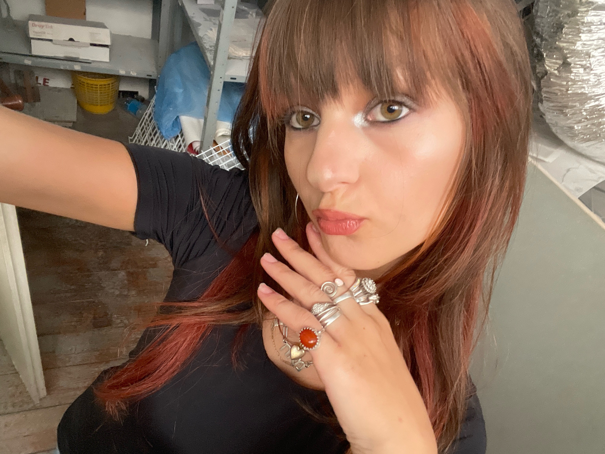 A young woman with reddish-brown hair and bangs, wearing a black t-shirt, takes a selfie in what appears to be a storage area or workshop. She is adorned with several silver rings on her fingers, one of which features a red stone and a heart charm. Her makeup includes noticeable eyeliner and lip gloss. The background is cluttered with shelves holding various items, including boxes, a yellow basket, and blue plastic sheeting. A metal duct is visible on the right side of the image. The lighting suggests an indoor environment, possibly with artificial illumination. The woman's pose and expression suggest a casual and informal moment captured for social media. There are no discernible elements that specifically identify the location as Raanana, Israel.