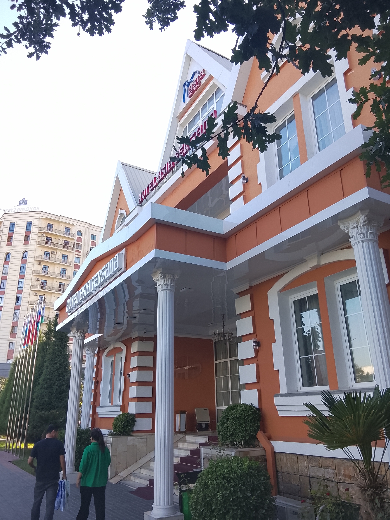 The image shows the Hotel Asia Fergana, located in Fergana City, Uzbekistan. The hotel is an orange building with white trim, featuring large windows and ornate columns. In front of the hotel, there are several flags and lush green trees. Two individuals, a man and a woman, are walking on a paved pathway. In the background, a taller, light-colored building is visible. The sky is bright and overcast.
