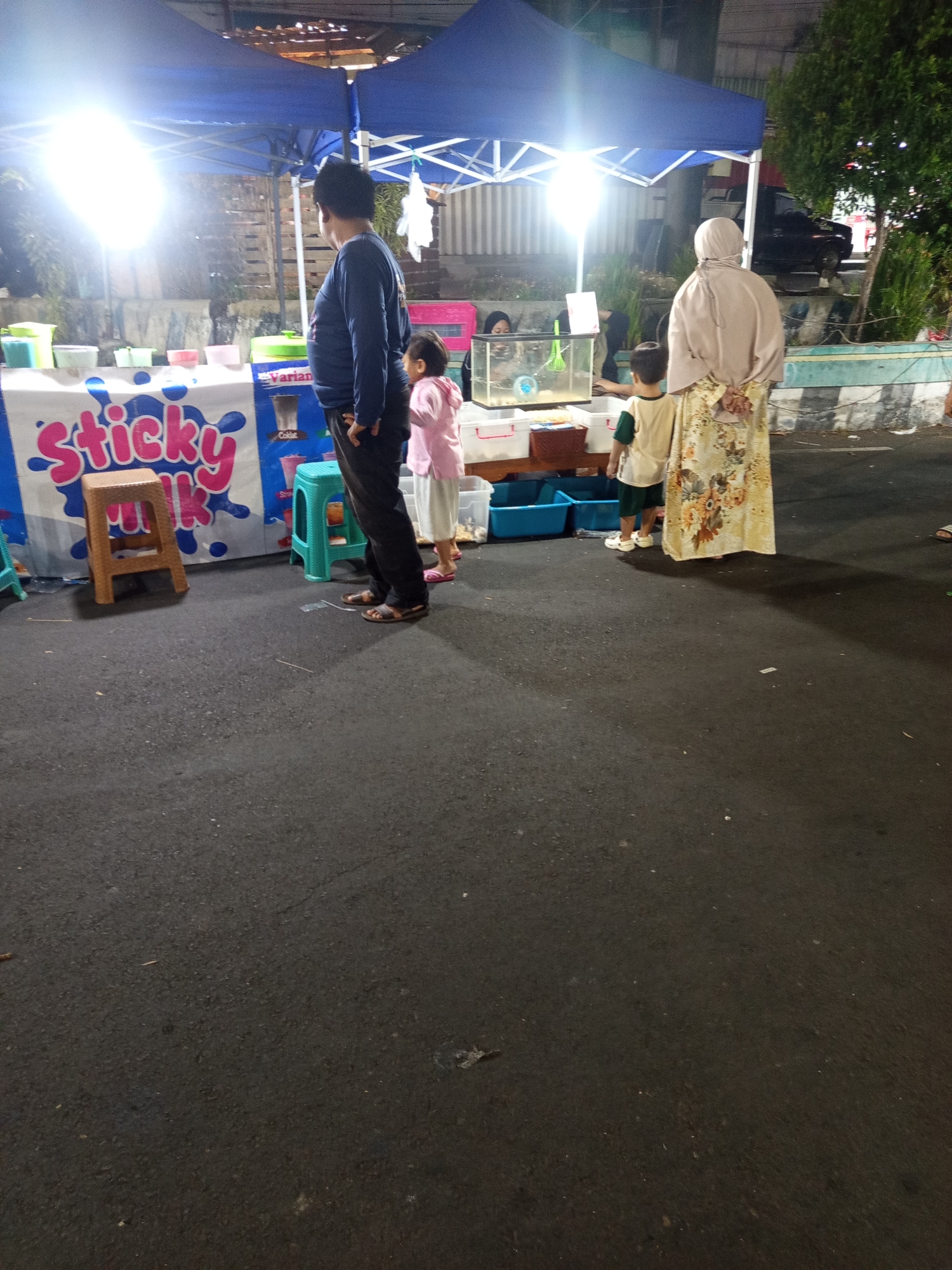 At dusk in Ardirejo, Indonesia, a scene unfolds at a street-side vendor. A person, likely an adult, stands facing away from the camera, wearing a dark long-sleeved shirt and dark pants. To their left, a young child in a pink shirt and white pants looks towards the vendor's stall. Further to the right, an adult in a light-colored hijab and a floral-patterned dress stands with their hands behind their back, observing. A young child in a green and beige outfit stands in front of them, looking at the stall.
The stall itself is adorned with a blue and white banner that reads "sticky milk." Behind the banner, various containers and what appears to be a small aquarium are visible. A bright light illuminates the area from the vendor's tent. In the background, trees and a parked vehicle can be seen, suggesting an urban or semi-urban setting. The ground is paved with asphalt, indicative of a street or market area. The overall atmosphere is that of an evening street market.
