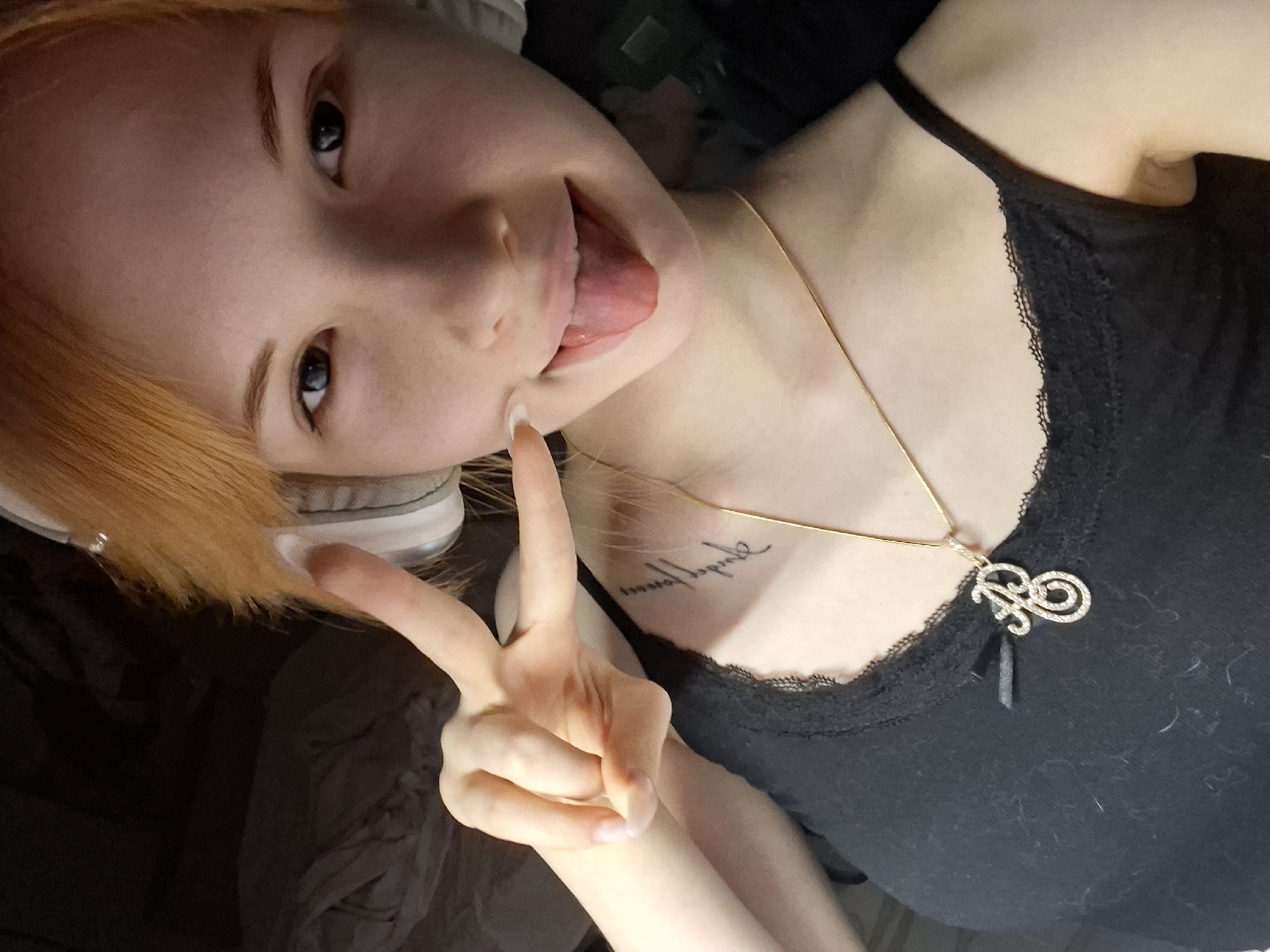 A young person with fair skin and vibrant orange-blonde hair is depicted in a close-up, upward-angled shot. Their blue-grey eyes are focused on the camera, and they are playfully sticking out their tongue. They wear a black tank top with a delicate lace trim along the neckline. A thin gold chain necklace with a sparkling, stylized initial pendant, appearing to be a 'B', adorns their chest. On their upper left chest/shoulder area, a tattoo with unreadable cursive script is visible.
The subject's right hand is raised, making a peace sign, with long, light-colored nails visible. Grey headphones are partially visible, resting on the left side of their head amidst their hair. The background is dark and indistinct, suggesting an indoor setting without revealing specific details. The lighting is soft and ambient. The overall impression is casual and cheerful. While the location context of Chaikovsky, Russia, is provided, there are no visual cues within the image itself to confirm this specific geographical location.