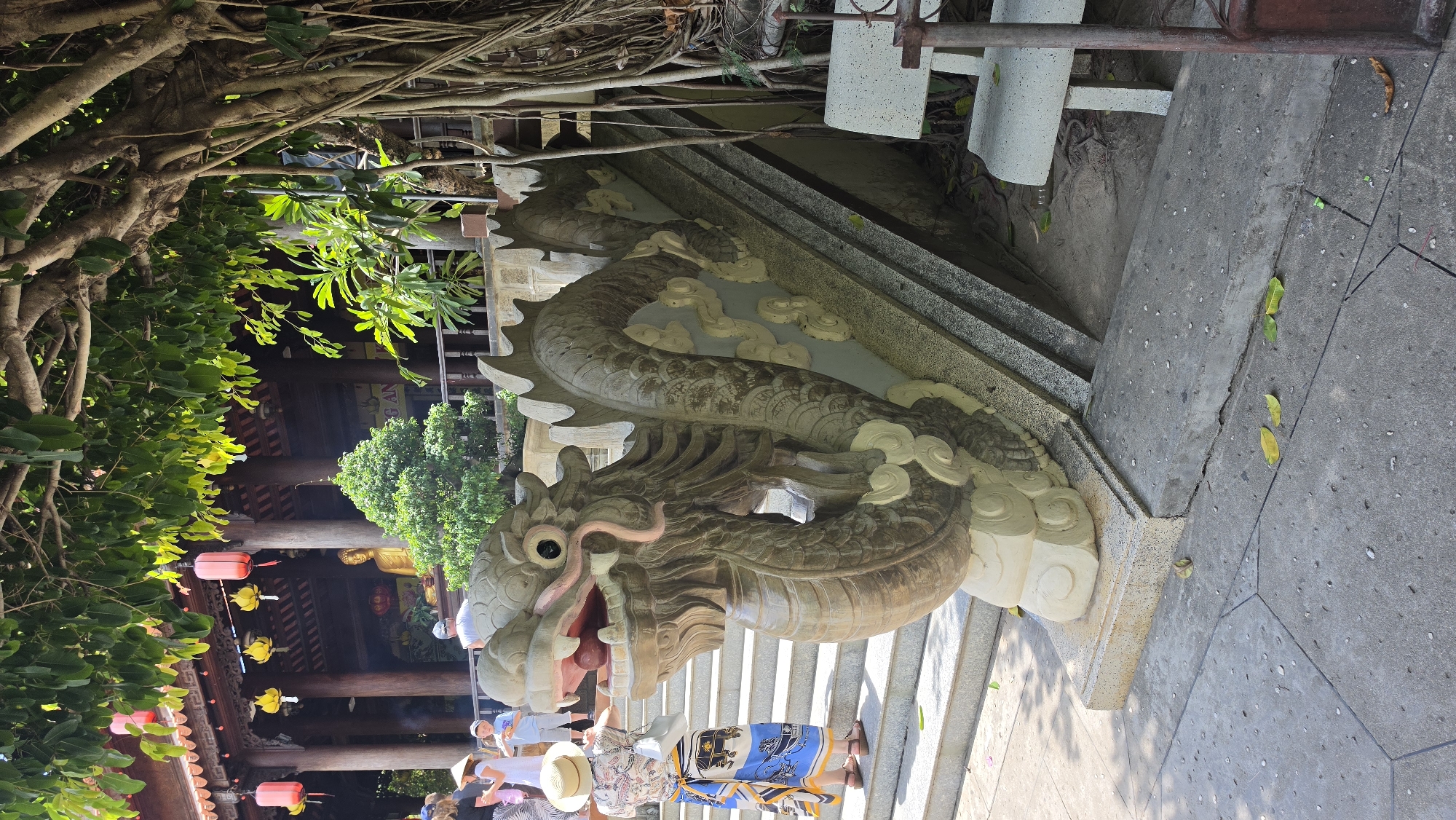 In Nha Trang, Vietnam, a weathered stone dragon sculpture serves as a decorative balustrade on the stairs of a temple. The dragon, depicted with an open mouth and a pink tongue, curves along the side of the steps. Behind the sculpture, people are visible walking up the stairs towards the temple building, which features traditional Vietnamese architecture with wooden pillars and a tiled roof adorned with red lanterns and yellow floral decorations. A golden Buddha statue is situated within the temple's interior. The scene is set outdoors under the shade of a large tree with exposed roots. In the foreground, the concrete ground is scattered with fallen leaves. A white stone bench is placed to the right of the stairs.